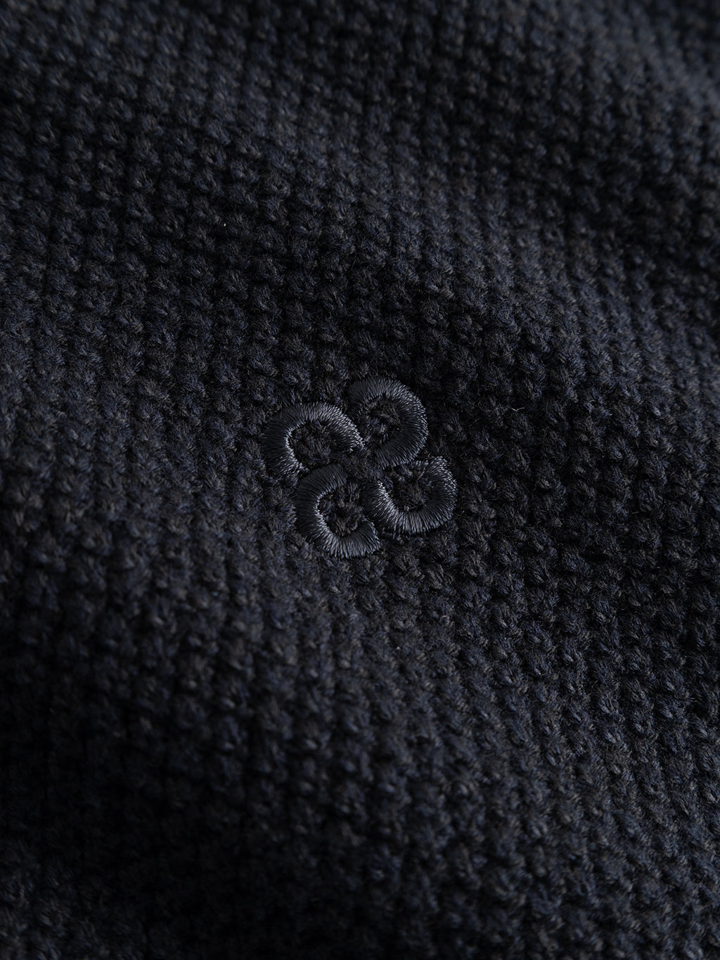 Close-up of the GABlamix Coro O-Neck LS Knit by Gabba in Blue Black, featuring a fine cotton-blend knit fabric and a subtly embroidered clover-shaped logo in matching thread at the center—highlighting the refined detailing characteristic of this classic crew neck sweater.