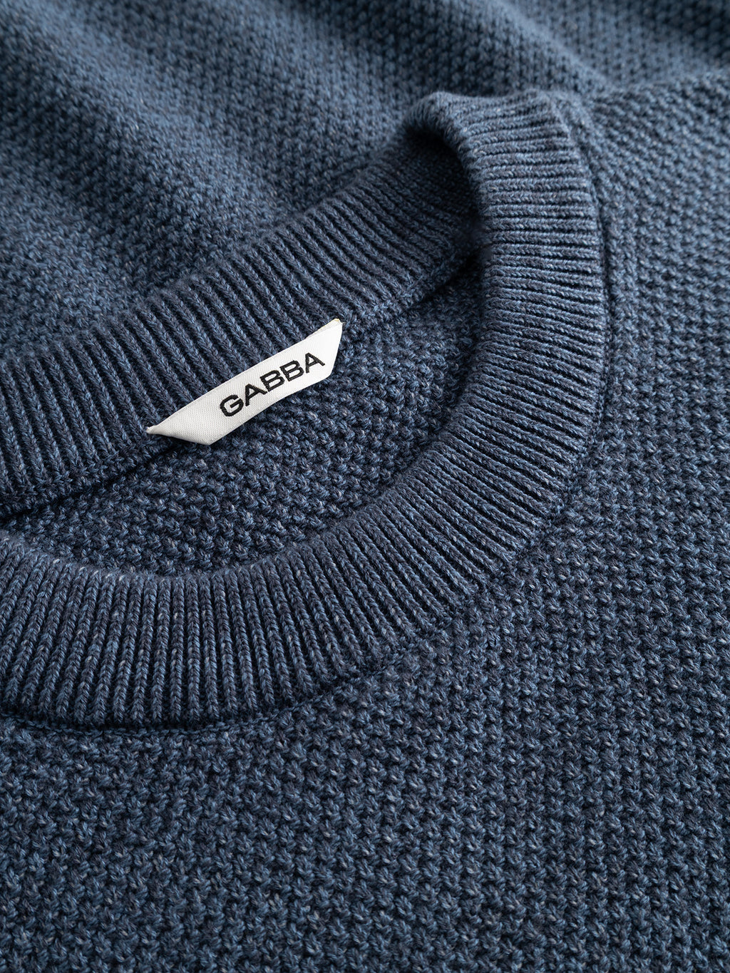 Close-up of the GABlamix Coro O-Neck LS Knit sweater by Gabba in Turbulence Blue, featuring textured dark blue cotton-blend fabric, a ribbed crew neck, and a white Gabba label attached near the collar.