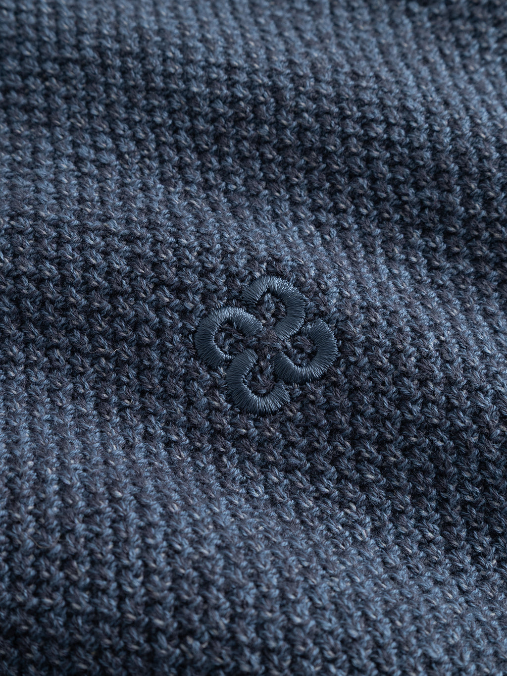 Close-up of the Gabba GABlamix Coro O-Neck LS Knit in Turbulence Blue, showing its textured dark blue cotton-blend fabric and a small, embroidered circular floral pattern in matching thread at the center.