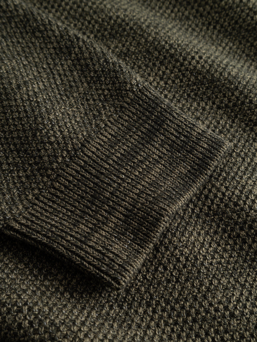 Close-up of the GABlamix Coro Half LS Knit in Forest Night by Gabba, featuring a dark-colored, textured cotton-blend sleeve resting on a matching knit surface. The ribbed cuff is clearly visible, highlighting the fabric’s detailed stitching pattern and emphasizing its regular fit.