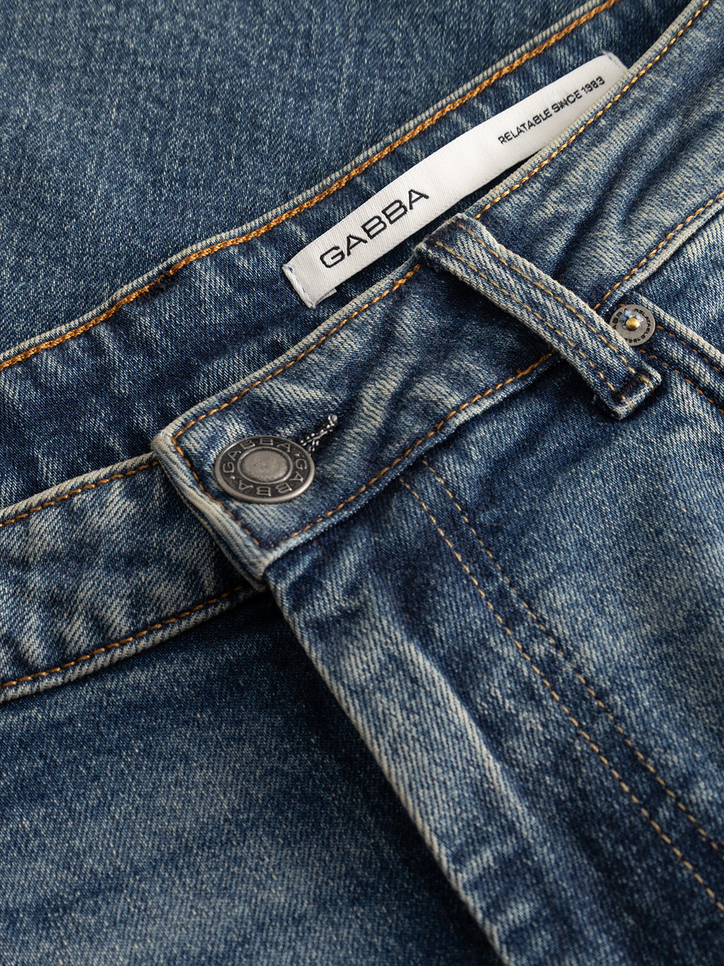 Close-up of the GABmarc Silvi DS0007 Jeans in mid blue denim by Gabba, featuring a regular fit crafted from comfort stretch fabric, visible stitching, metal button, belt loops, and a white label inside the waistband that reads GAËBA RELIABLE SINCE 1983.