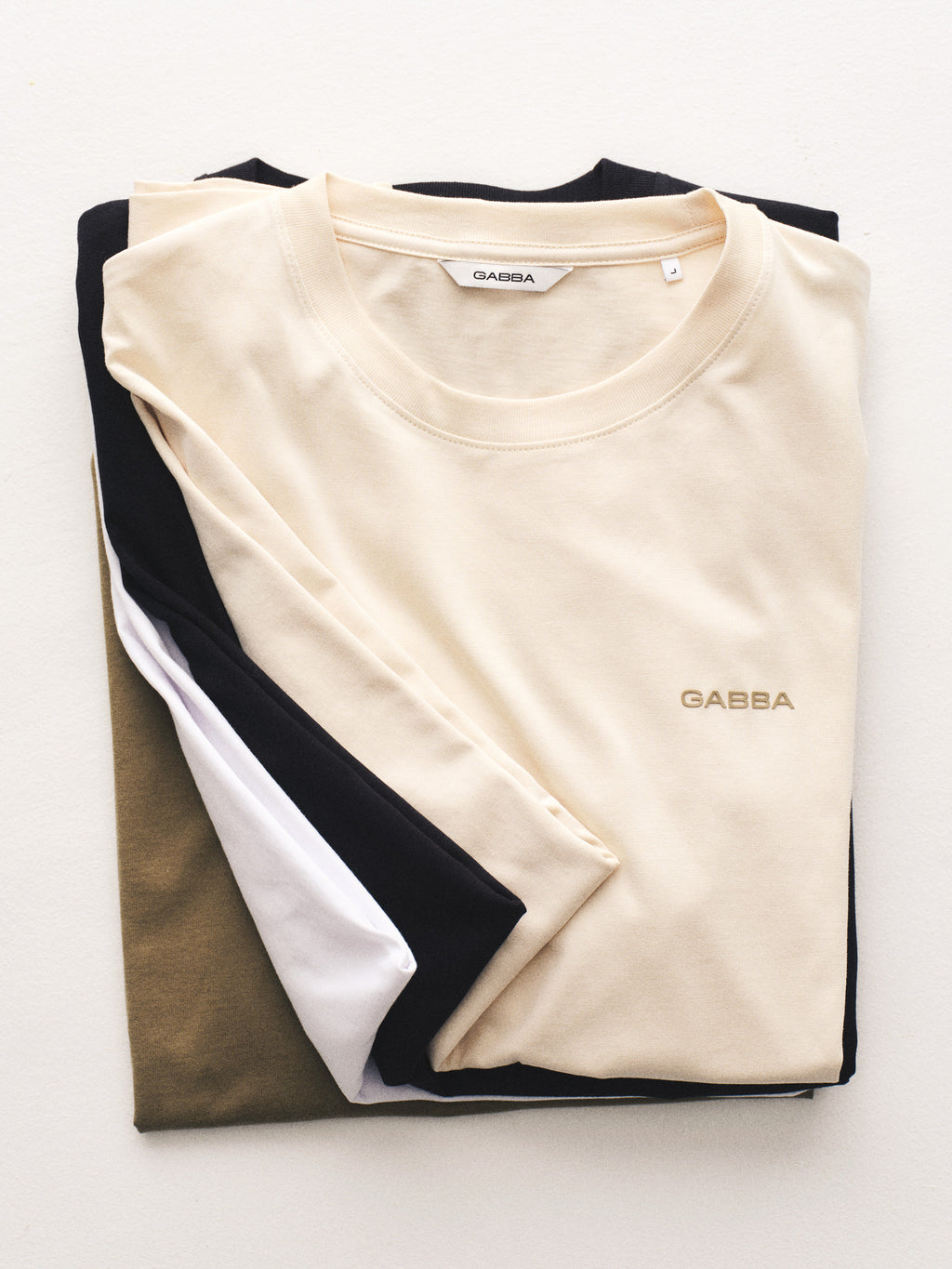 A neatly folded stack of four classic fit T-shirts in beige, black, white, and olive green, with the top shirt—the Gabba GABdune Logo GOTS SS T-Shirt in Birch—showing the Gabba logo on the chest.