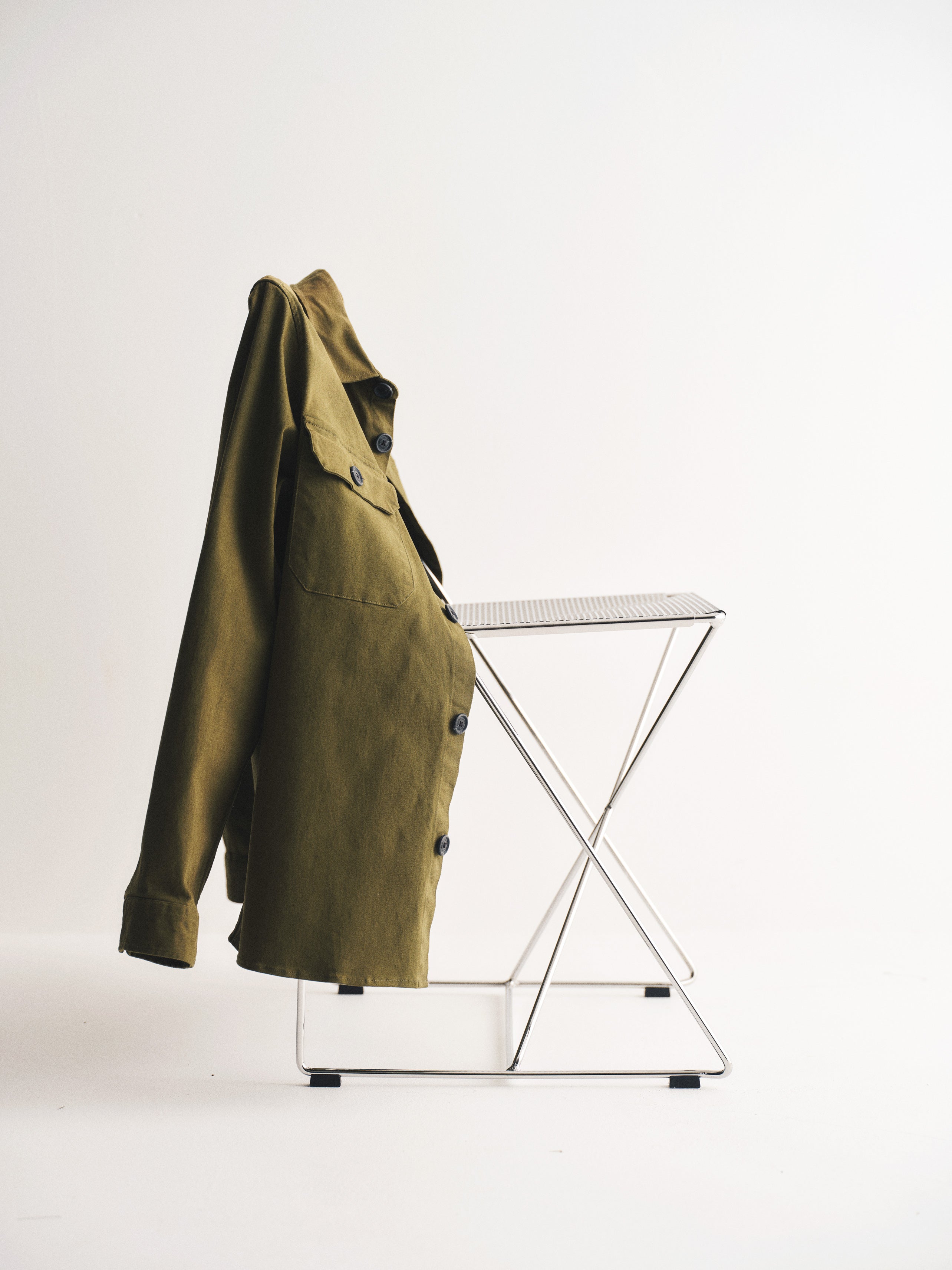 The GABBA GABtopper LS Shirt in Army, a heavyweight overshirt jacket, hangs neatly over the back of a minimalist metal folding chair set against a plain white background.