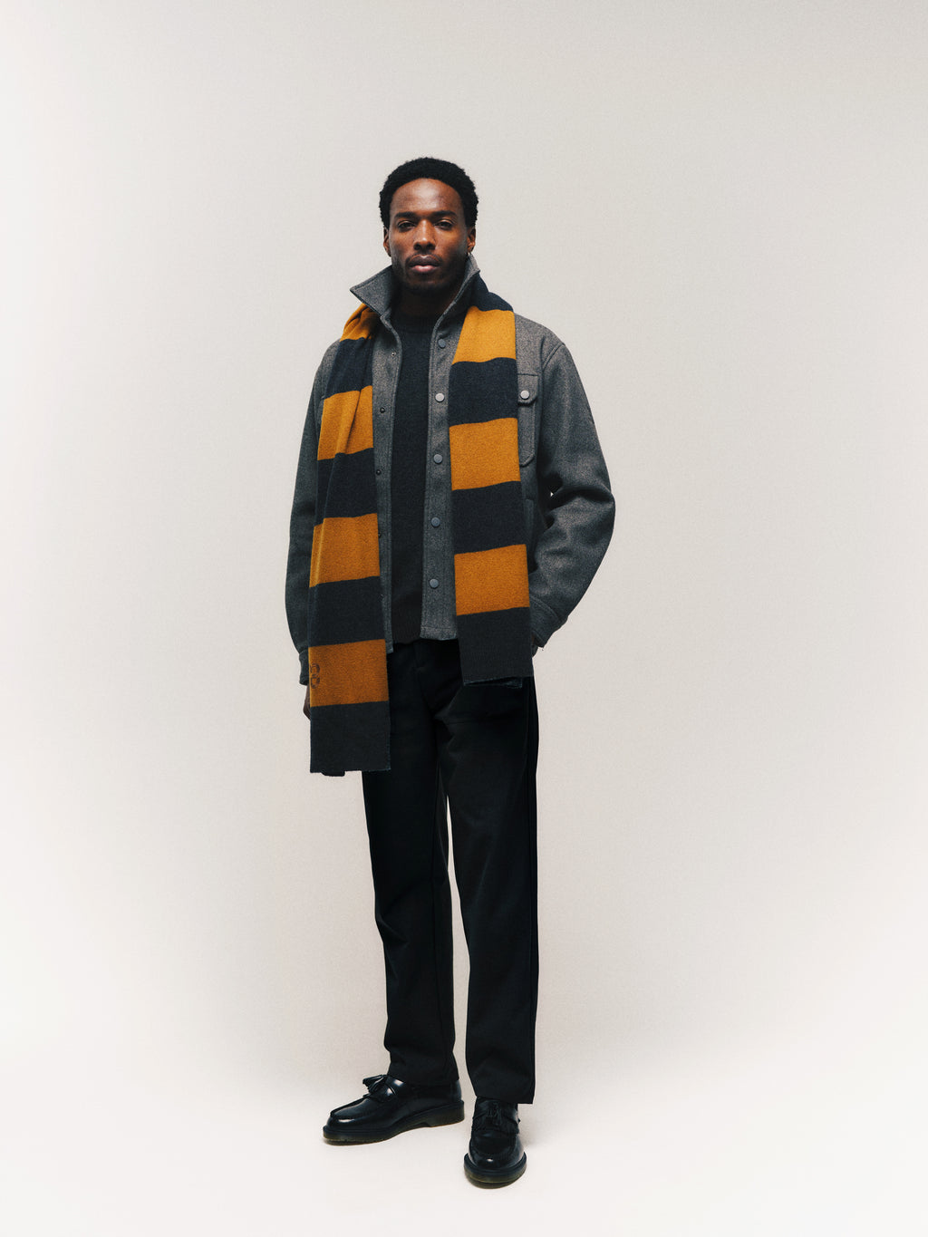 A man stands against a plain background, wearing the Gabba GABtheo City Jacket in dark grey, perfect for everyday layering, paired with dark pants, black shoes, and a yellow and black striped scarf.