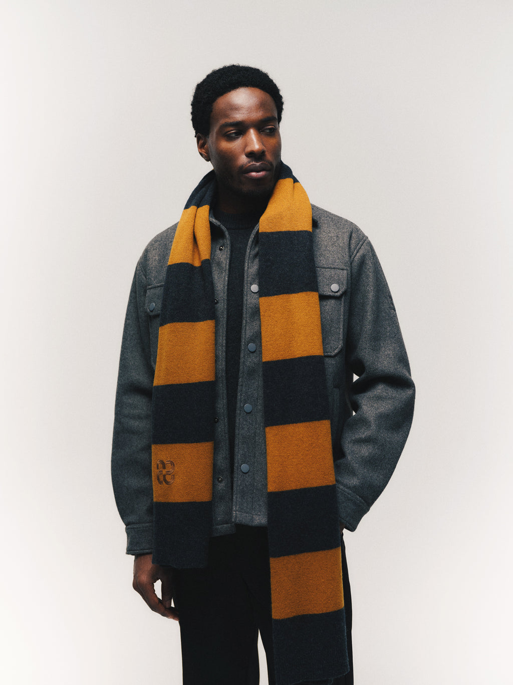 A man wearing the Gabba GABtheo City Jacket in dark grey over a dark shirt, with a long yellow and black striped scarf draped around his neck, stands against a plain, light background—perfect for everyday layering.