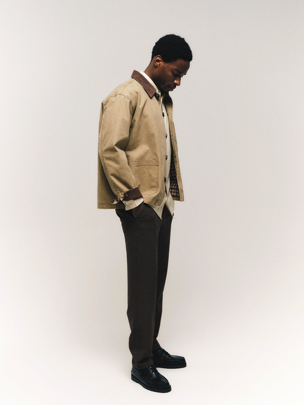 A man stands with his head slightly bowed, wearing the Gabba GABotto Tela Outerwear - Sand Crocker—a beige washed cotton canvas workwear jacket featuring a subtle corduroy collar—paired with brown trousers, a light shirt, and black shoes against a plain off-white background.
