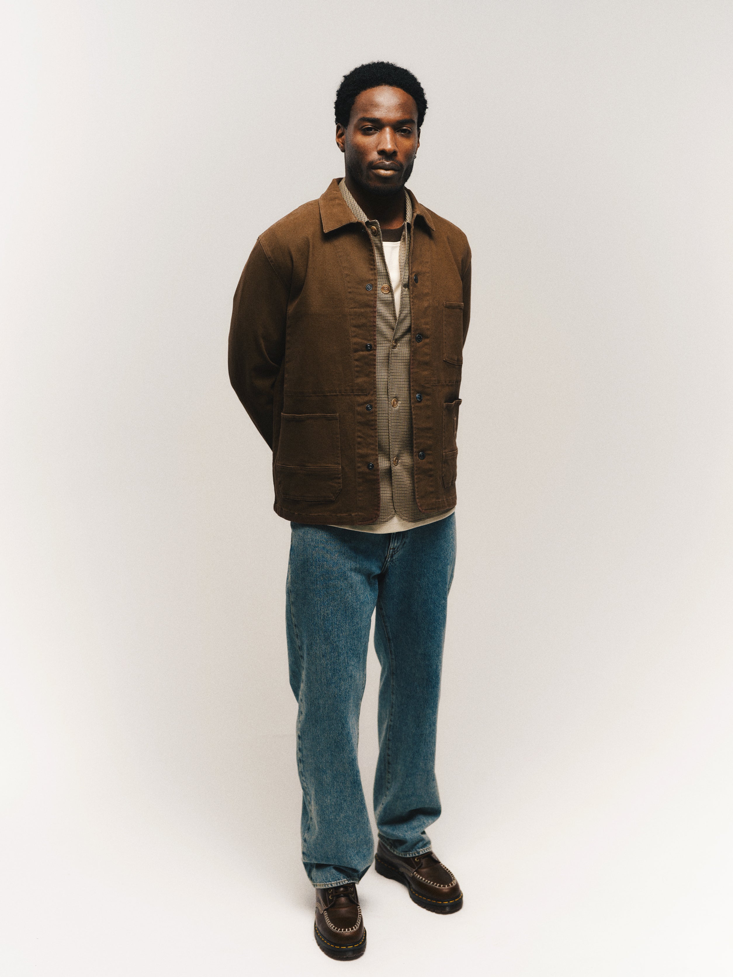 A man stands against a plain white background, wearing the Gabba GABbeta Twill - Brown Slate jacket over a tan shirt, regular fit blue jeans with utility pockets, and brown shoes, with his hands behind his back.