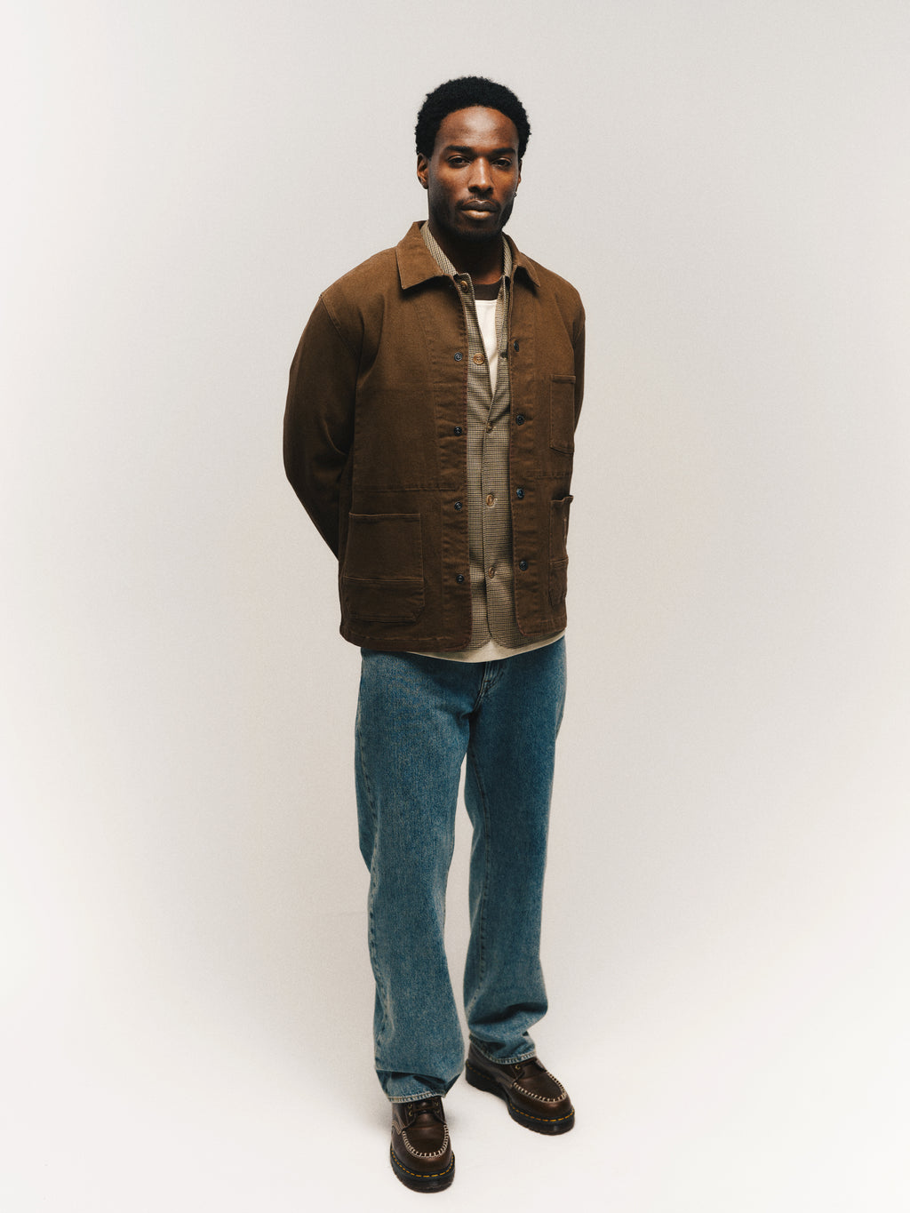 A man stands against a plain white background, wearing the Gabba GABbeta Twill - Brown Slate jacket over a tan shirt, regular fit blue jeans with utility pockets, and brown shoes, with his hands behind his back.