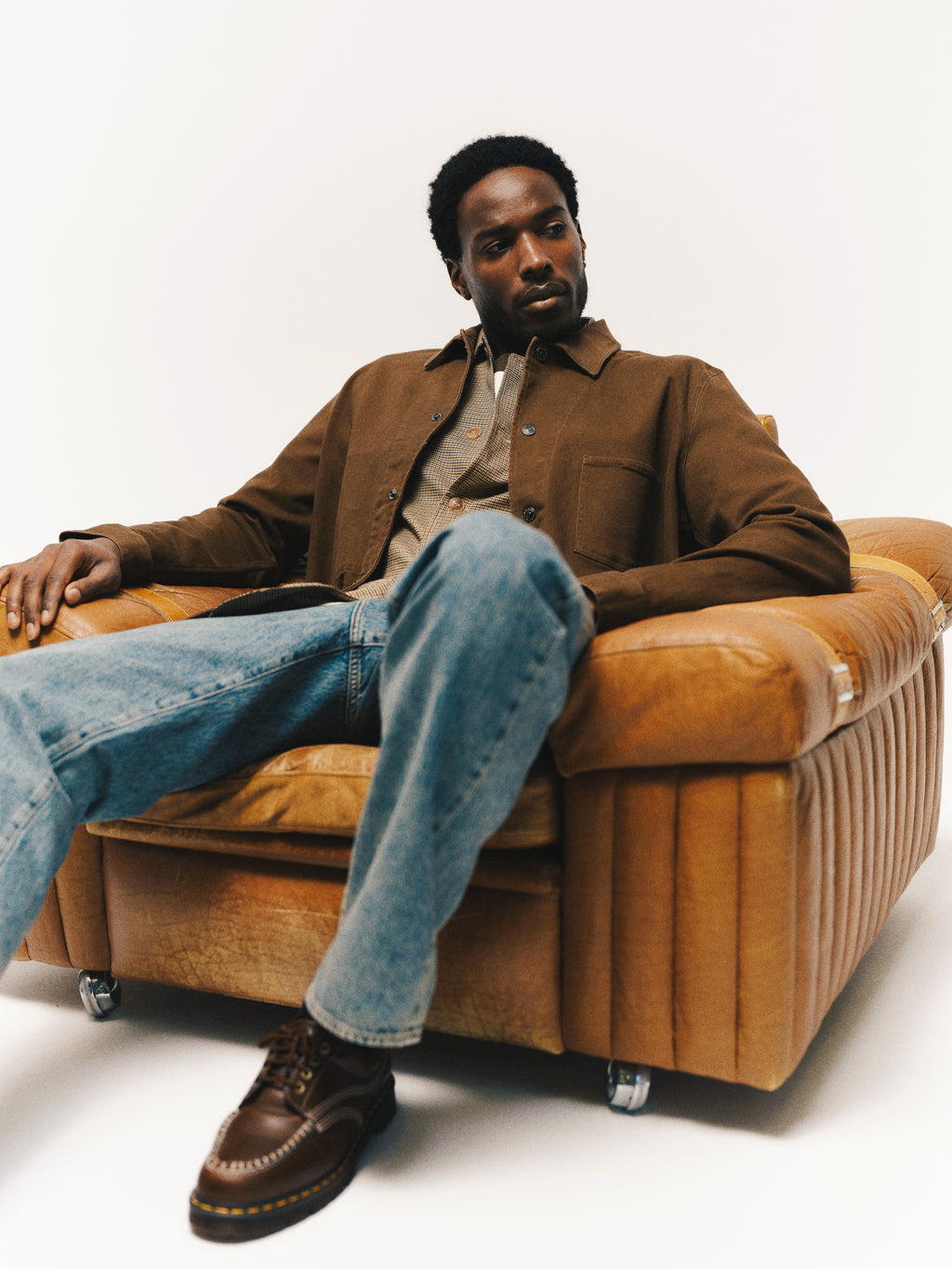 A man wearing a brown jacket, checkered shirt, and Gabba GABnoah Tona RS2100 Jeans in Mid Blue Denim made from organic cotton sits relaxed on a vintage tan leather armchair, looking to the side against a plain white background.
