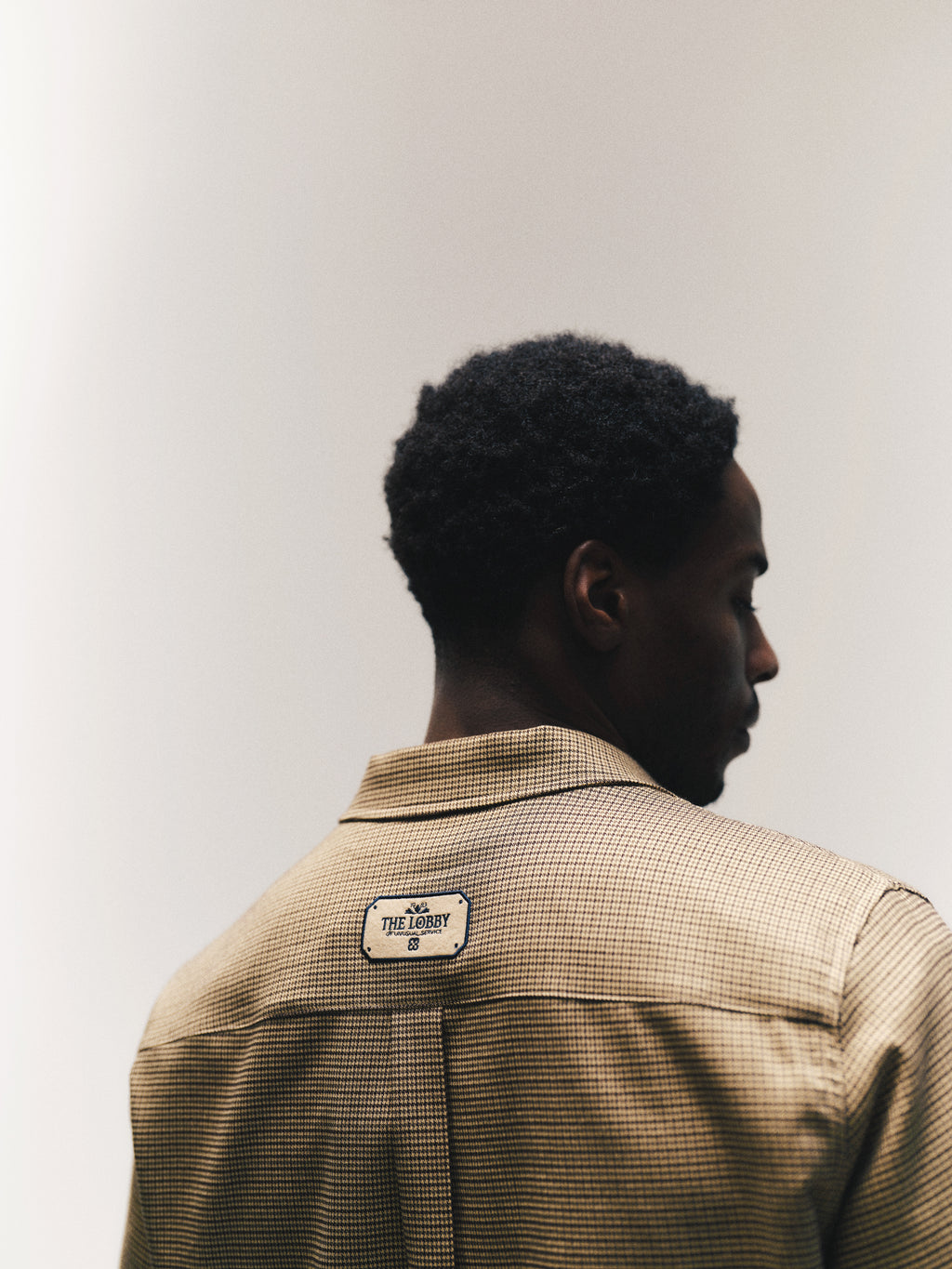 A man with short curly hair is facing away, wearing the Gabba GABfelix Brit LS Shirt in Dark Sand—a relaxed fit shirt featuring a brown checkered pattern and a label on the back that reads THE LOBBY. The background is plain and softly lit.