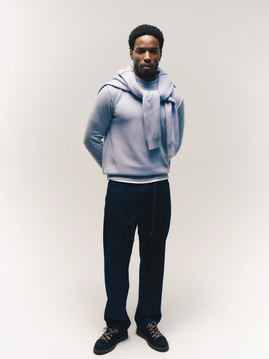 A man stands against a plain background, wearing a light blue sweater with another sweater draped over his shoulders, GABnoah Tona Rinse Jeans in dark blue denim by Gabba, and black shoes. His hands are behind his back as he looks calmly at the camera.