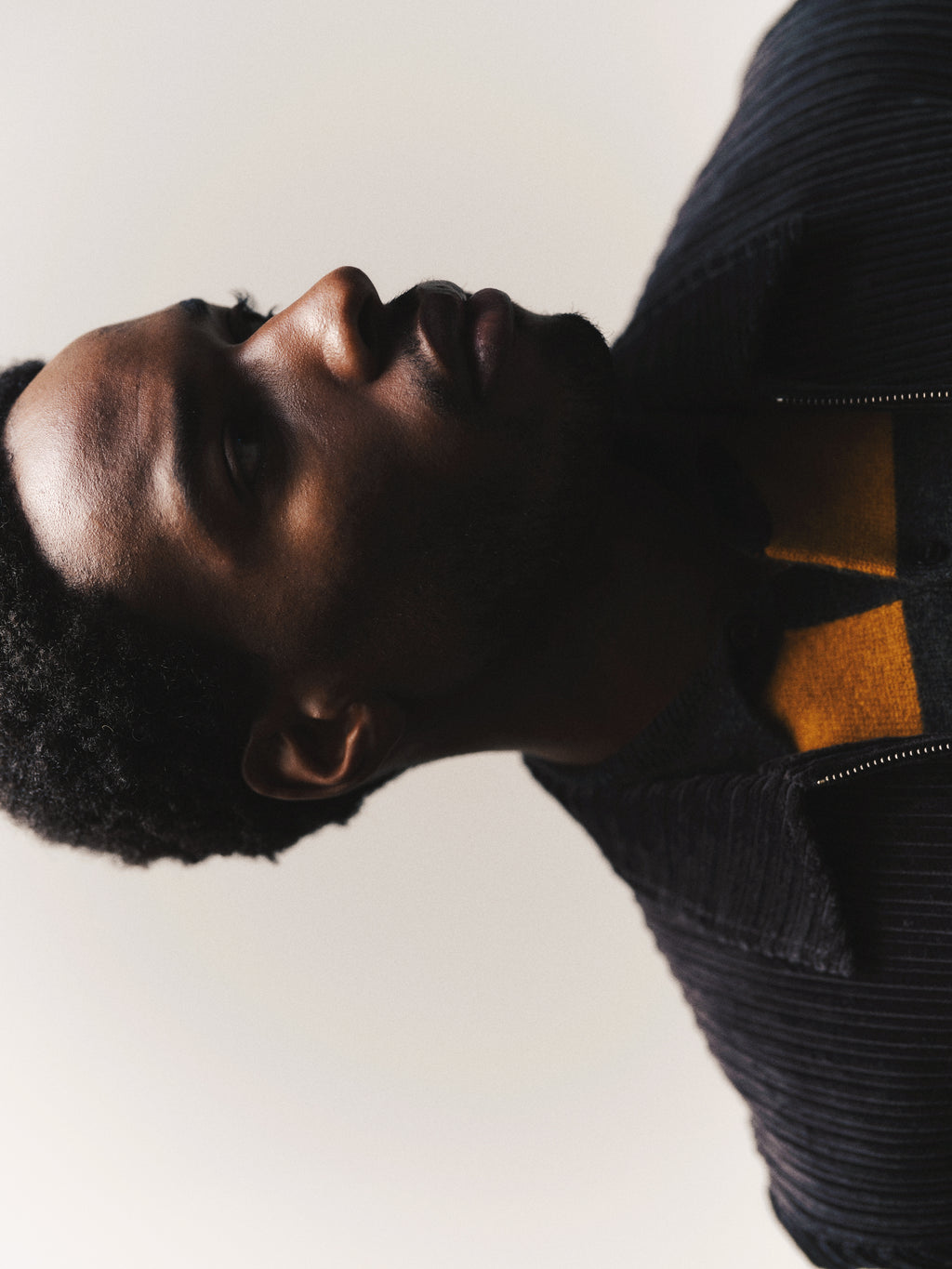 A man with short hair, dressed in a dark jacket layered over the Gabba GABworren Polo Stripes Aico LS Knit in yellow stripes (men’s regular fit), is photographed in soft light against a pale background, gazing slightly upward and to the side.