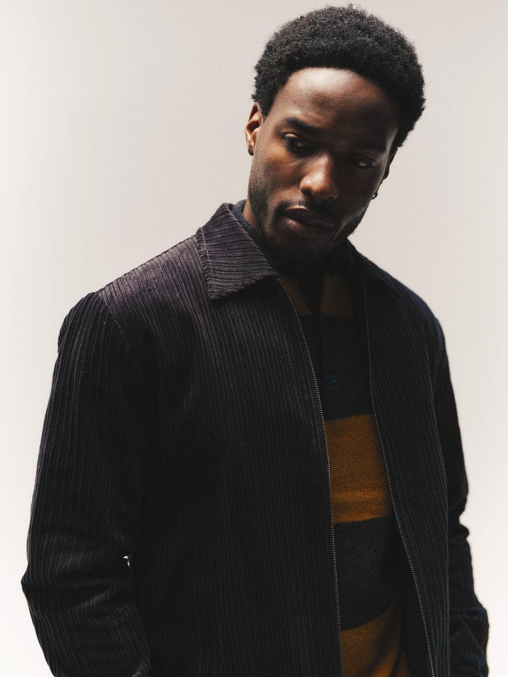 A man with short curly hair, dressed in a dark corduroy jacket layered over the Gabba GABworren Polo Stripes Aico LS Knit in yellow stripes, looks down with a neutral expression against a plain light background.