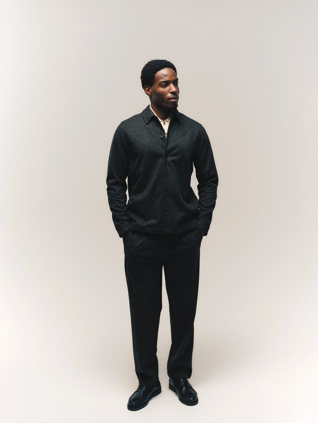 A man stands against a plain light background, gazing to his right. He wears a dark button-up shirt, black shoes, and Gabba’s GABnapoli 19 Zaffe GRS Pant in Dark Grey—a regular straight fit trouser with a wool-look finish—with his hands in his pockets.