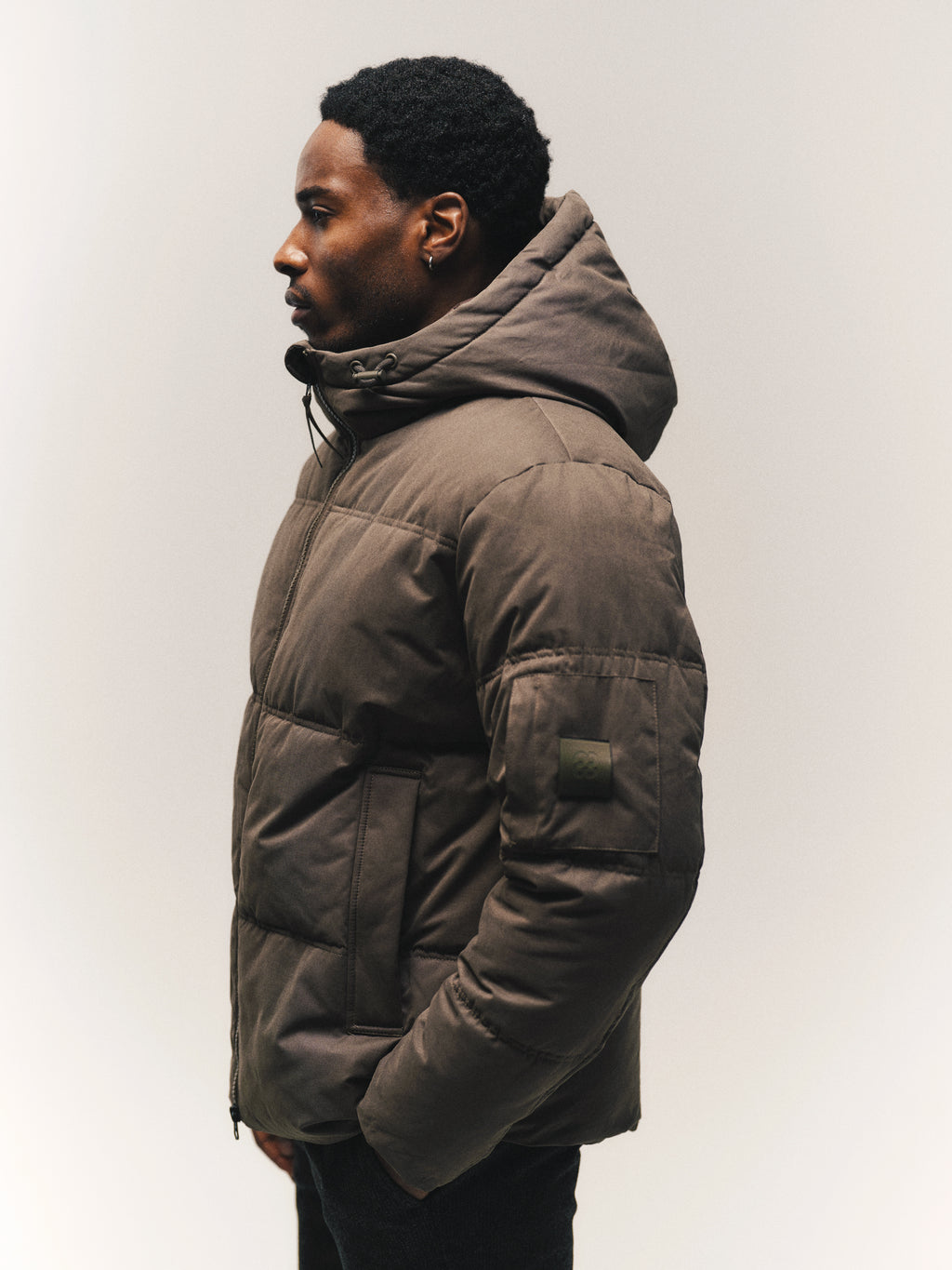 A man in profile wears a brown hooded GABjulien Calle Outerwear - Brown Slate puffer jacket with an adjustable hood, a zippered front, and a small patch on the sleeve.