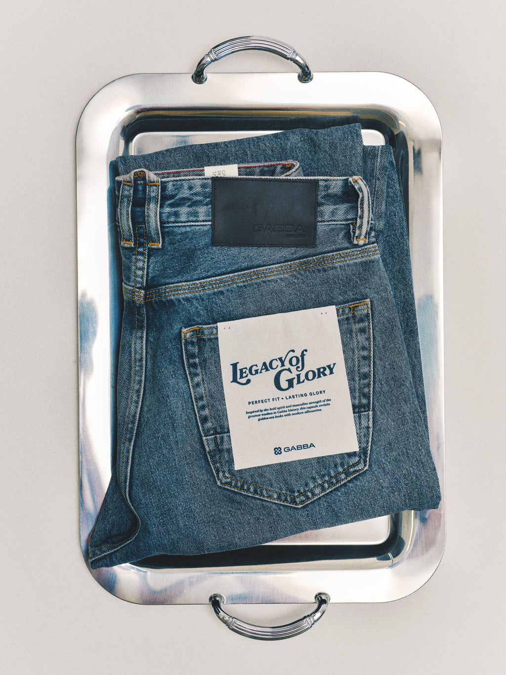 A pair of GABcarl Stone RS2103 Jeans in mid-blue denim by Gabba, made from premium ISKO fabric and adorned with the signature Legacy of Glory label, is neatly folded on a gleaming silver tray with handles set against a plain background.