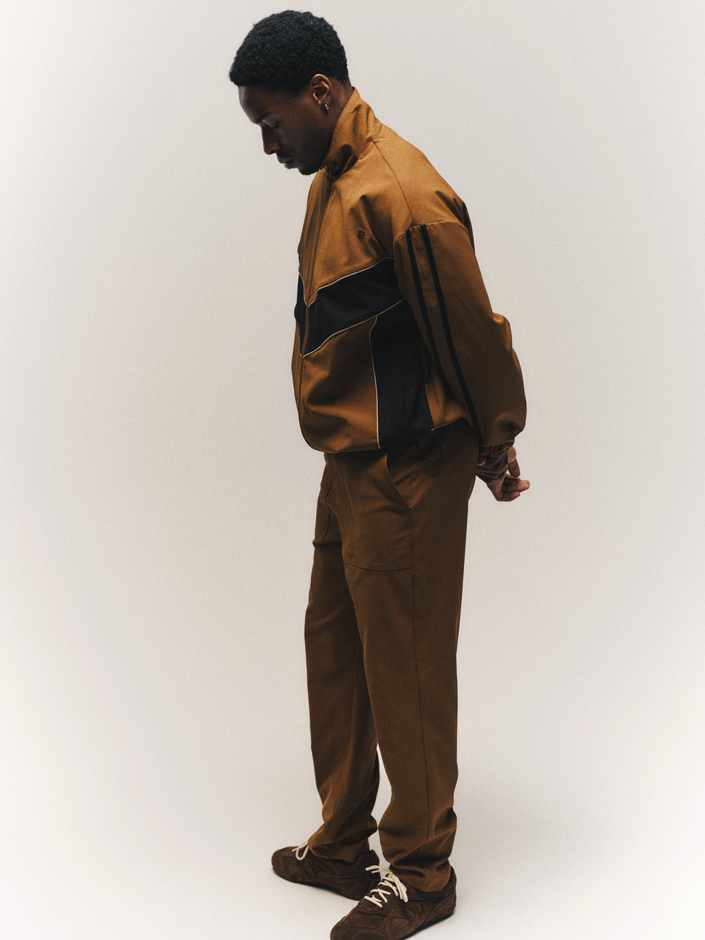 A man wearing the Gabba GABtrack Nord Jacket in brown, featuring black accents, stands sideways against a plain light background, looking down with his hands behind his back.