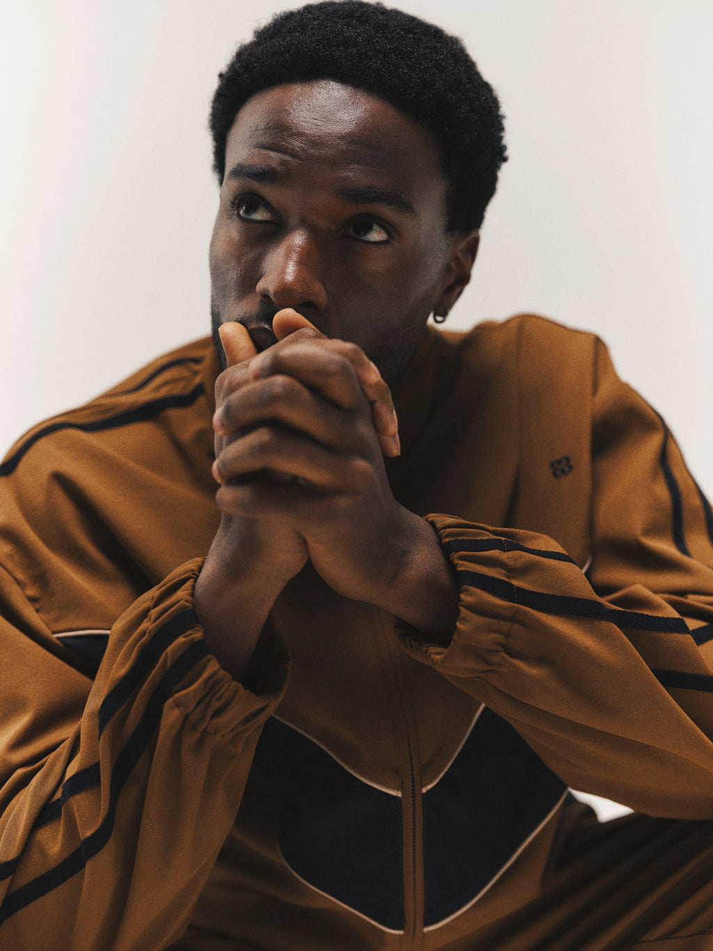 A person in the Gabba GABtrack Nord Jacket in brown sits with their hands clasped near their face, gazing upward in a thoughtful pose. The jacket’s relaxed fit enhances the sense of comfort, while the plain, well-lit background draws attention to their reflective mood.
