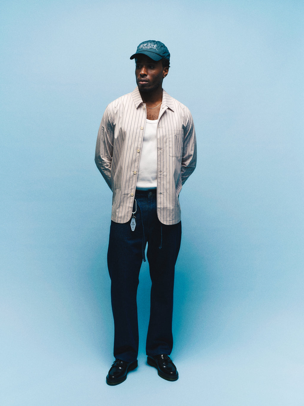 A man stands against a light blue background with his hands behind his back, wearing a blue cap and a striped shirt layered over a white tank top. He completes his effortlessly stylish look with Gabba’s GABnoah Tona Rinse Jeans in Dark Blue Denim—featuring a loose straight fit crafted from organic cotton denim for added comfort and sustainability.