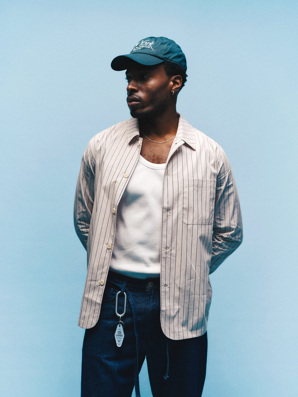 A man wearing a green cap, white tank top, striped shirt, and Gabba GABnoah Tona Rinse Jeans in dark blue denim stands against a light blue background, looking to the left with his hands behind his back.