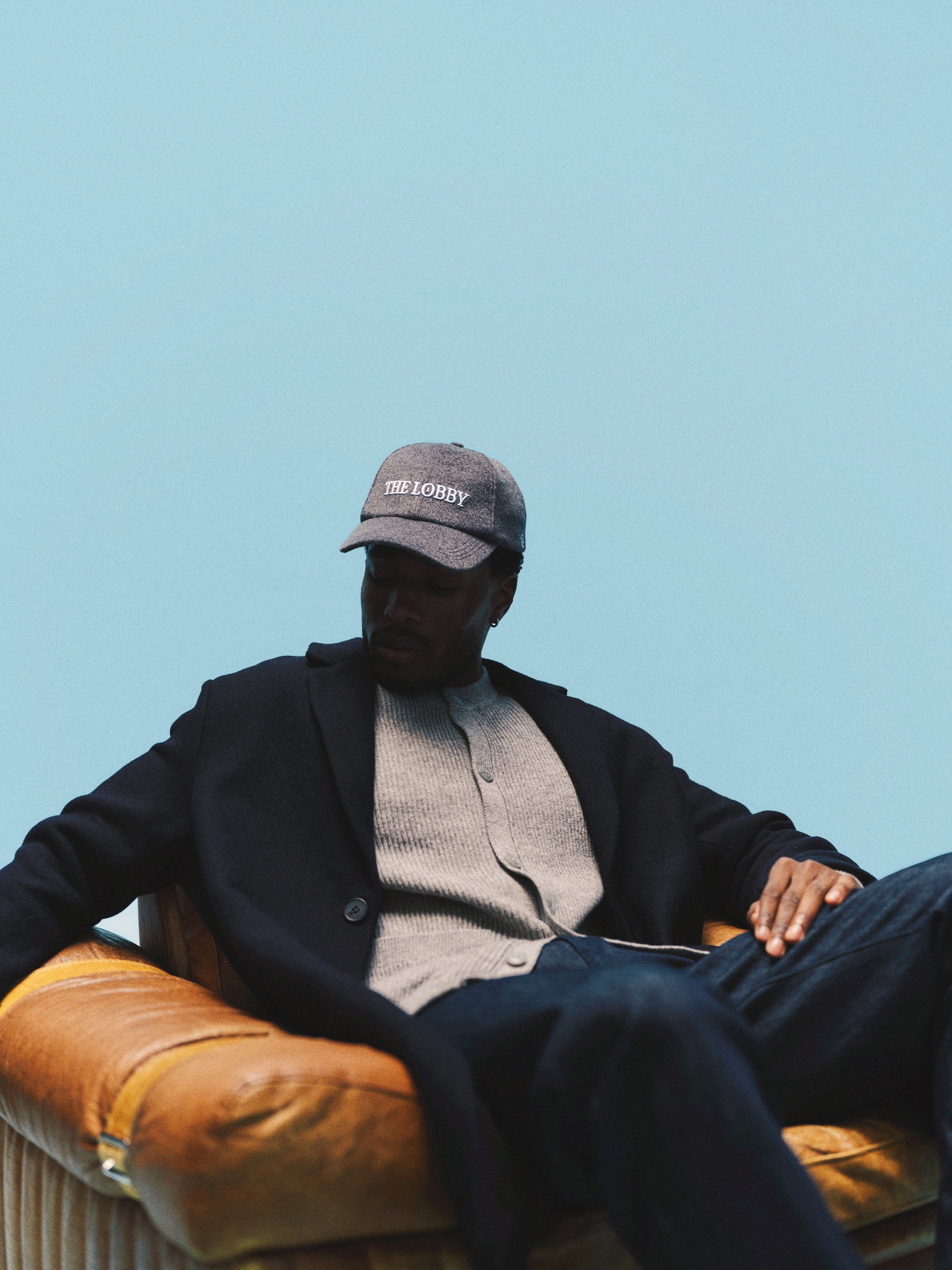A person in a dark coat and cap sits on a tan leather chair against a blue background, looking down with a relaxed posture, wearing Gabba’s GABcarl Anark Stone Raw Jeans in dark blue denim.