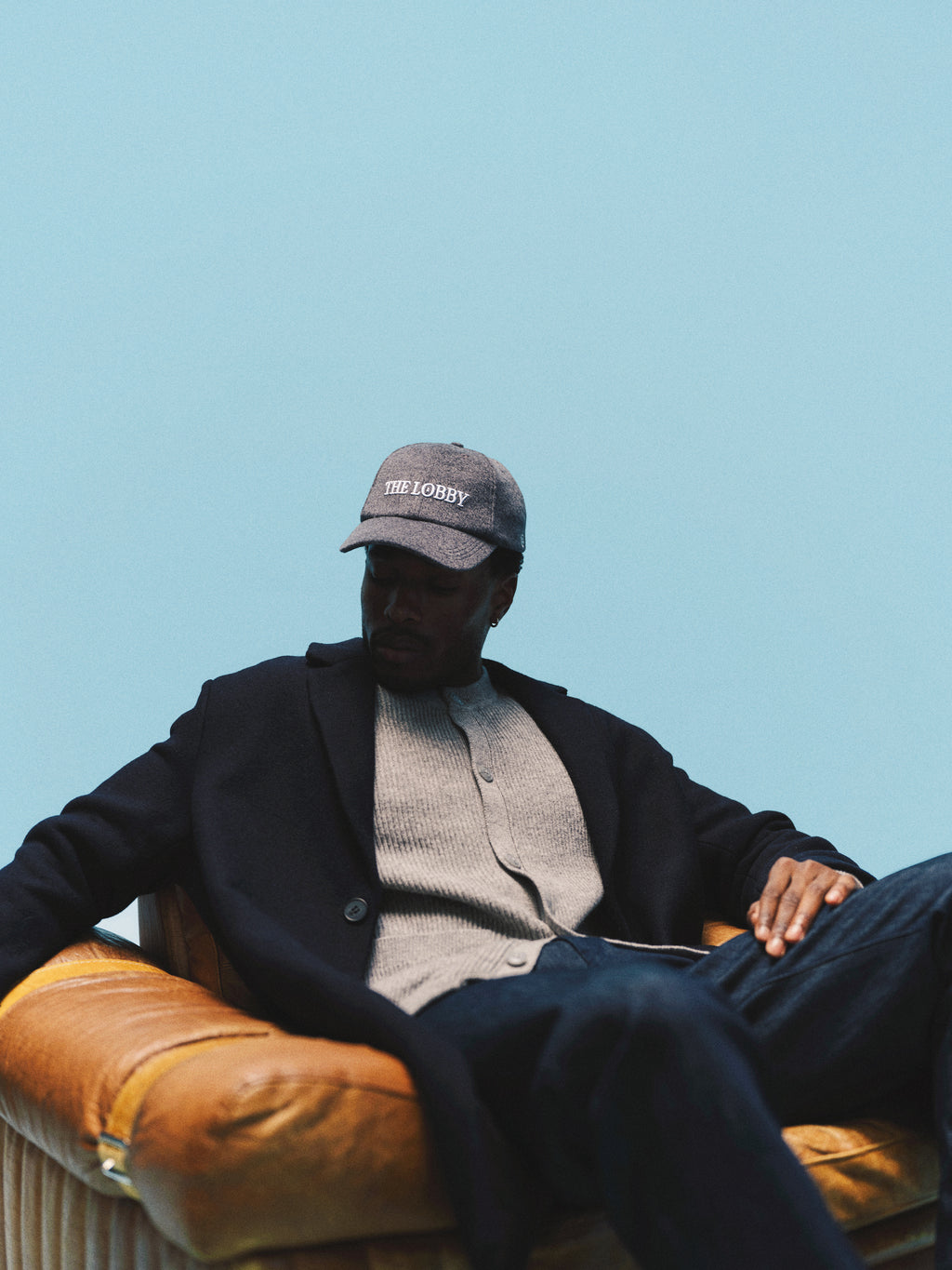 A person in a dark coat and cap sits on a tan leather chair against a blue background, looking down with a relaxed posture, wearing Gabba’s GABcarl Anark Stone Raw Jeans in dark blue denim.