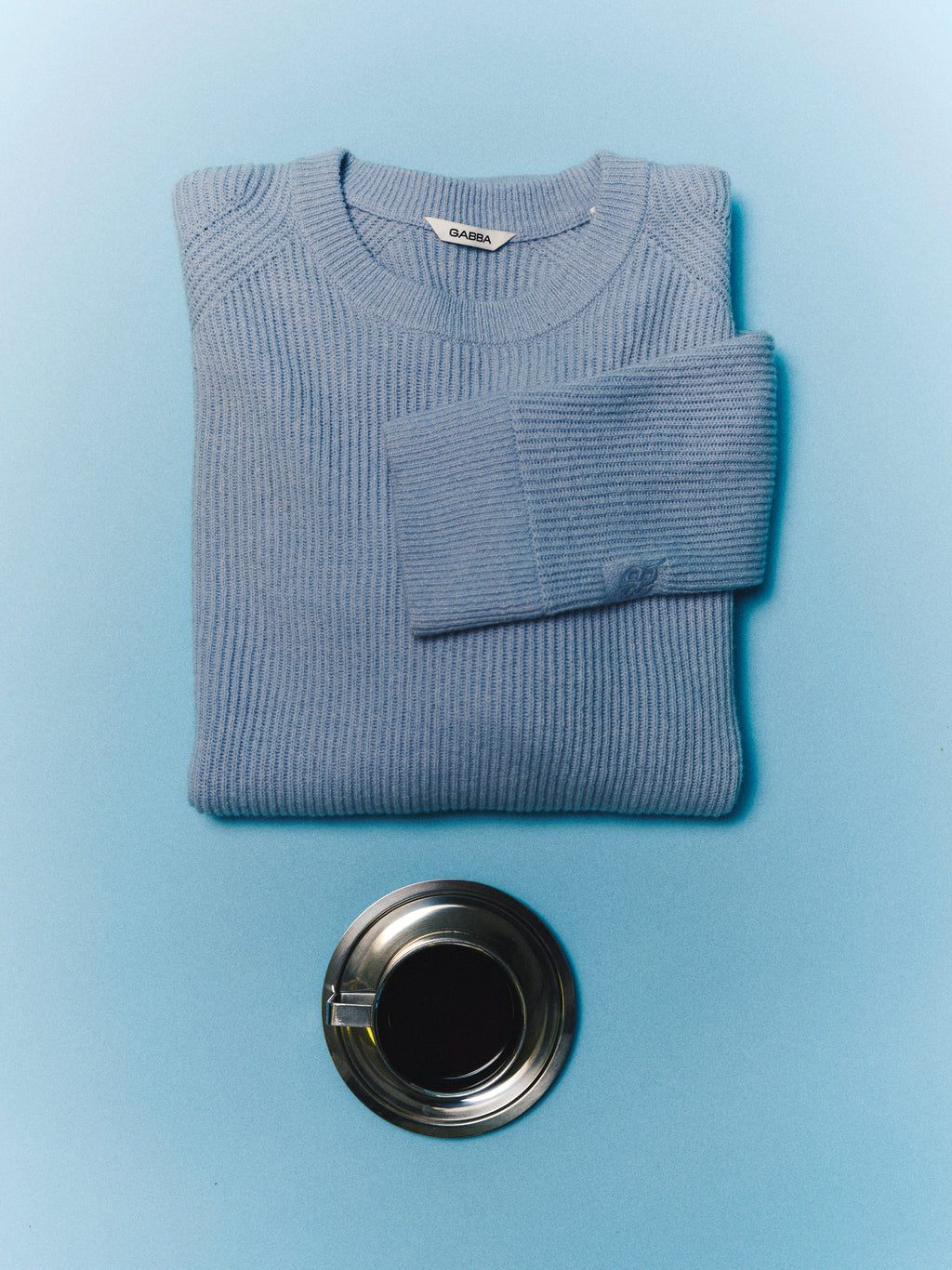 A folded Gabba GABremy O-Neck Flash LS Knit in Dusty Blue is placed above a full metal cup of black coffee, both set on a smooth, light blue surface.