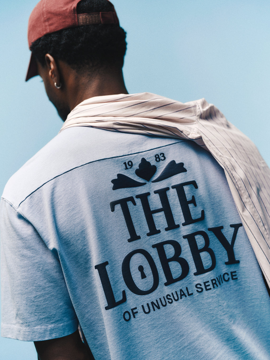 A person in a red cap and draped shirt faces away, wearing the Gabba GABlegin Wix Boxy Hour Lobby1 SS T-shirt in Dusty Blue—a relaxed boxy fit tee crafted from textured cotton jersey, featuring a bold back graphic with “THE LOBBY OF UNUSUAL SERVICE 1983” against a blue background.
