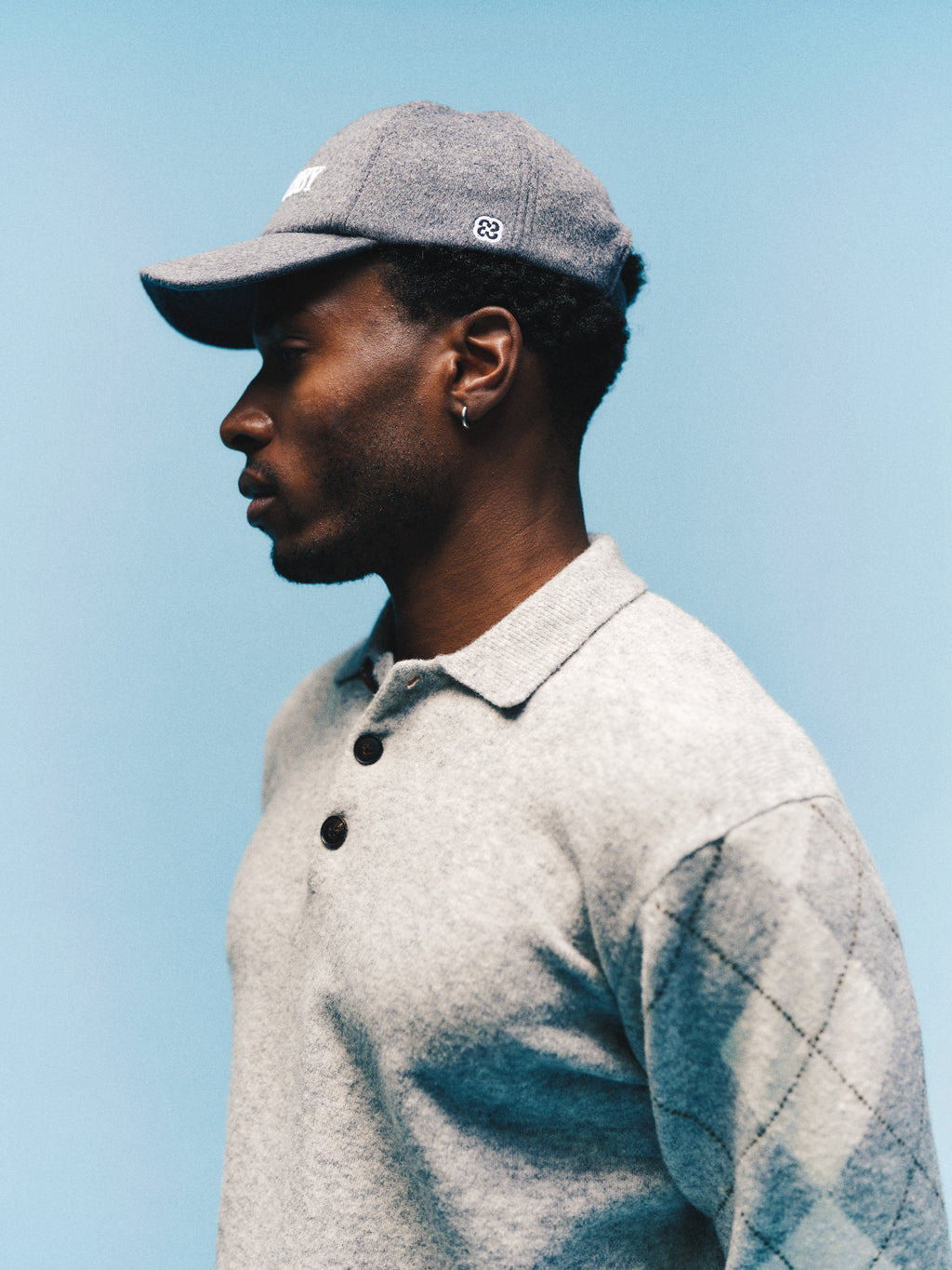A man wearing a gray cap and the Gabba GABpietro Polo Argyle LS Knit in Light Grey Mel, featuring modern diamond-patterned sleeves, stands in profile against a blue background.