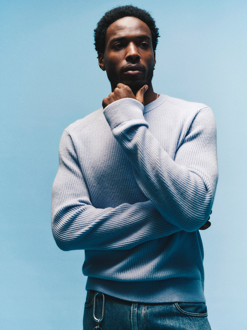 A man wearing the Gabba GABremy O-Neck Flash LS Knit in Dusty Blue and jeans stands against a blue background, one hand resting on his chin as he appears deep in thought. The relaxed-fit sweater, crafted from 100% wool, brings both comfort and contemporary style to his contemplative pose.