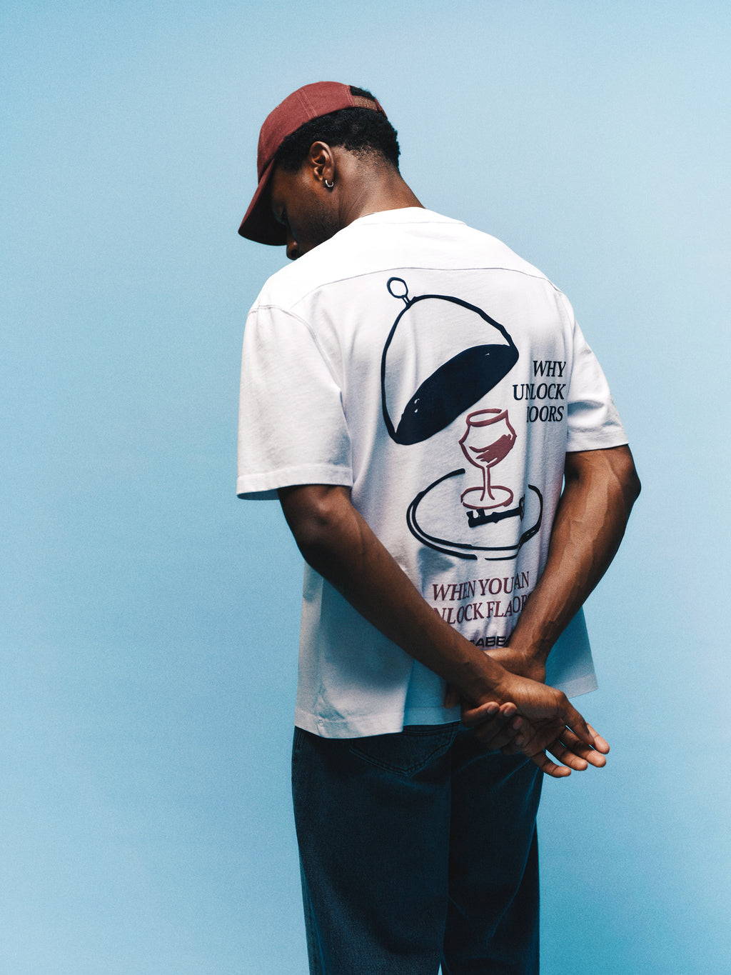 A person wearing the Gabba GABlegin Wix Boxy Hour Lobby2 SS T-shirt in white, featuring a relaxed boxy fit and graphic back print with text, paired with blue jeans and a maroon cap, stands against a blue background with their hands clasped behind their back.