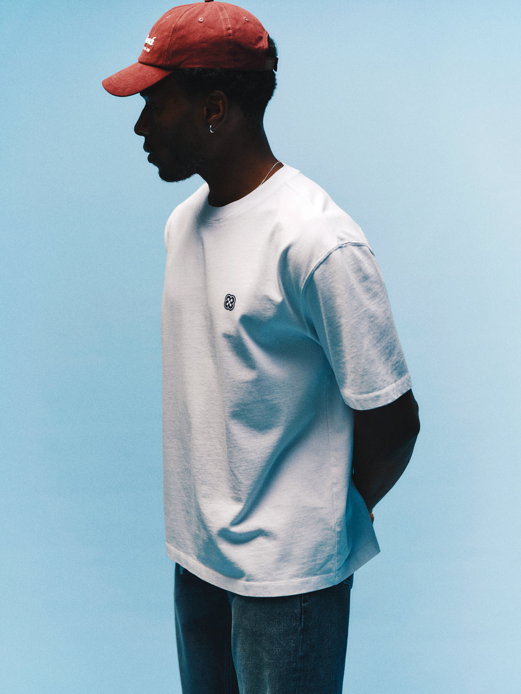 Here’s a revised sentence using your product data: A person in a red cap, Gabba GABlegin Wix Boxy Hour Lobby2 SS T-shirt in white, and jeans stands against a light blue background, facing left with hands behind their back.