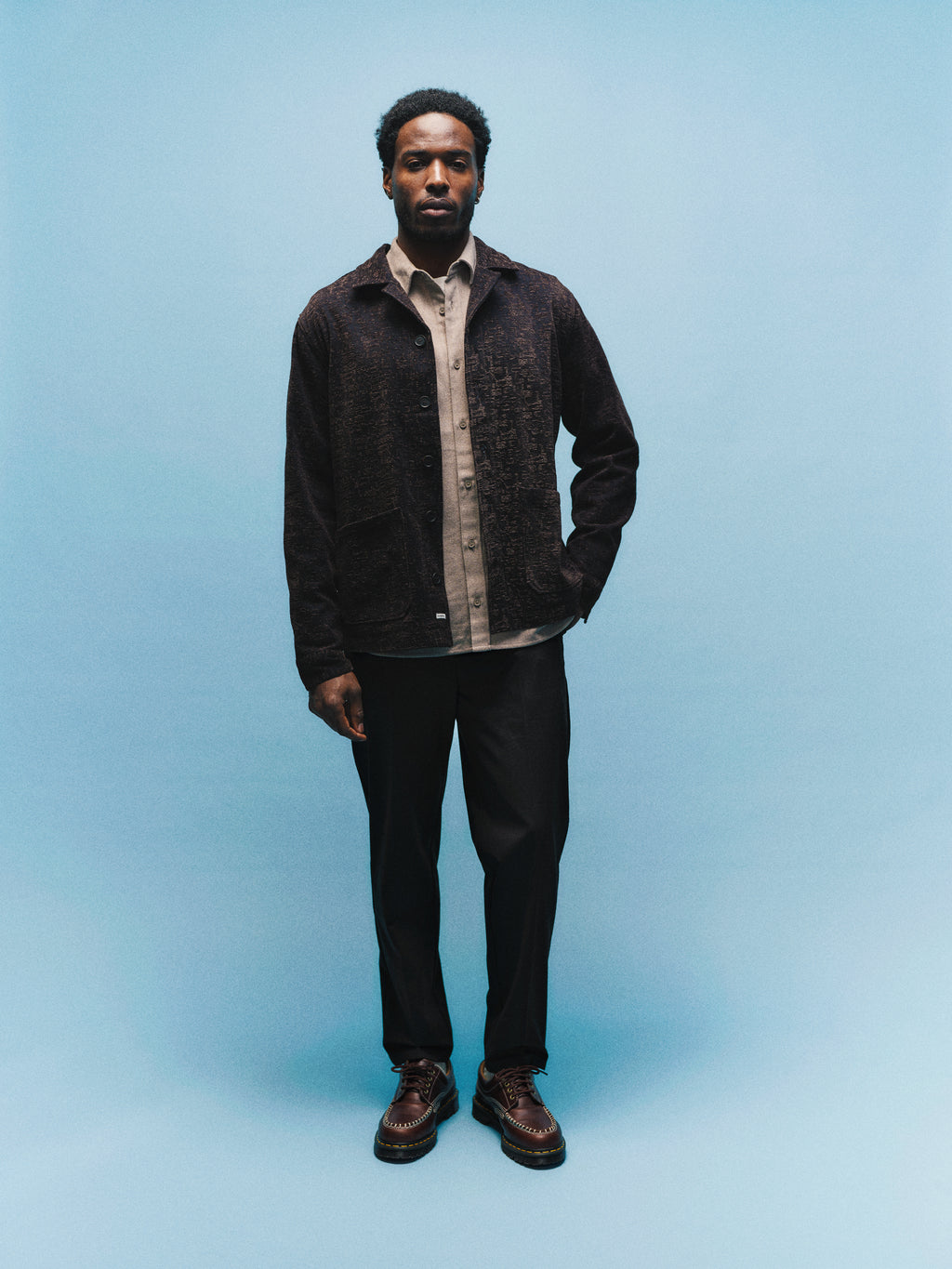 A man stands against a light blue background, wearing the Gabba GABsimone Pockt Monte LS Overshirt in Brown Slate, paired with black pants and brown shoes, looking straight ahead with a neutral expression.
