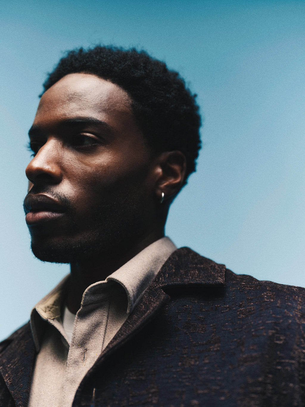 A man with short curly hair and an earring gazes to the side, wearing the Gabba GABsimone Pockt Monte LS Overshirt in Brown Slate, set against a blue gradient background.