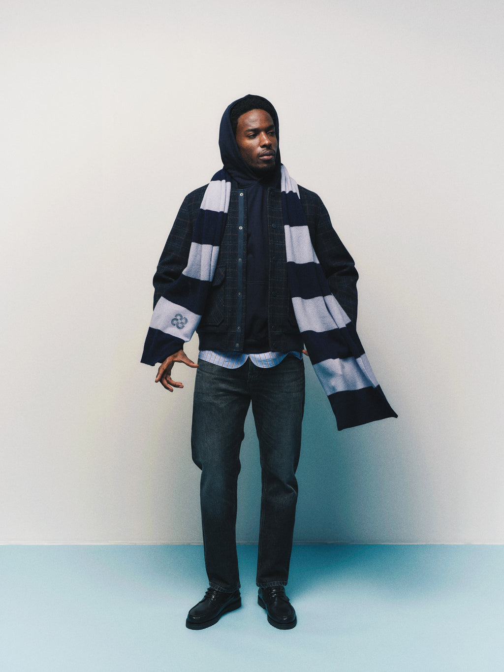 A man wearing the Gabba GABmatheo Check Bomber Jacket in blue check as a versatile outer layer, paired with a blue and white striped scarf, jeans, black shoes, and a subtly checked striped shirt, stands against a plain light background.