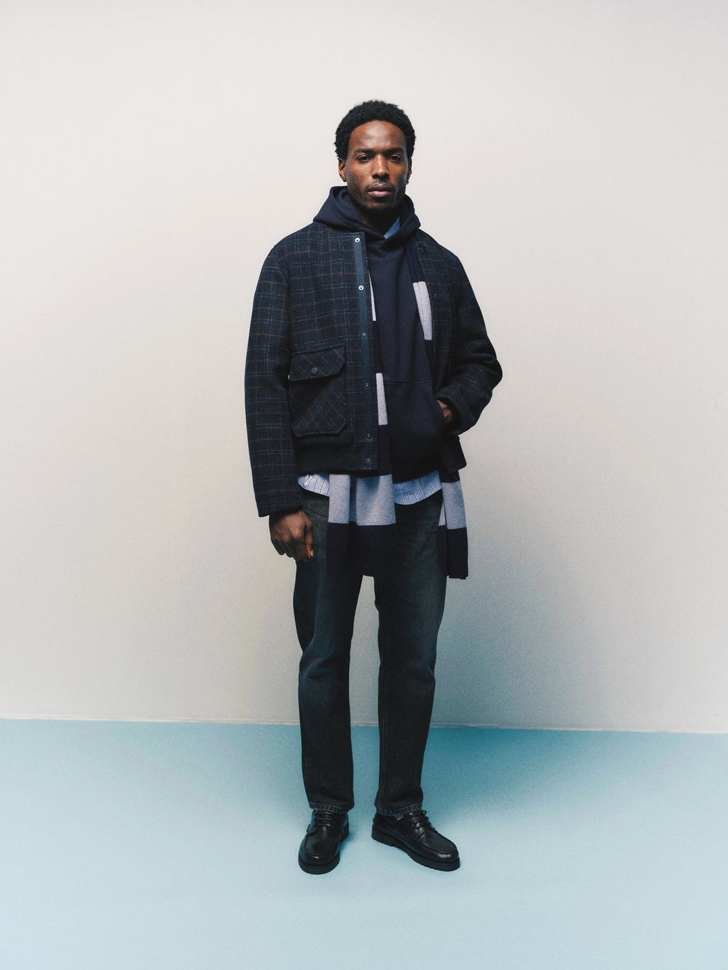A man stands against a light background wearing a dark plaid jacket, a navy hoodie, layered shirts, loose tapered fit Gabba GABcarl Blackrow DH0011 Jeans in black denim, and black shoes. He looks straight ahead with a neutral expression.