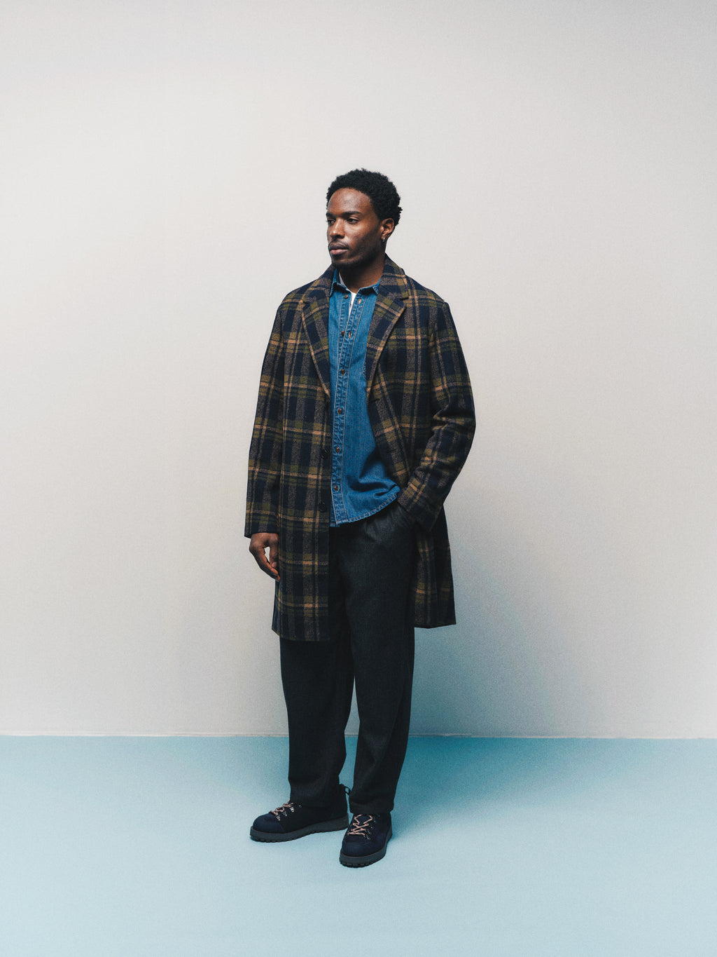 A man stands against a plain background wearing the Gabba GABsven Reg Wish LS Shirt in mid blue denim, paired with a plaid overcoat, dark trousers, and black shoes. He looks to the side with his hands by his sides, highlighting this versatile piece from Gabba’s collection.