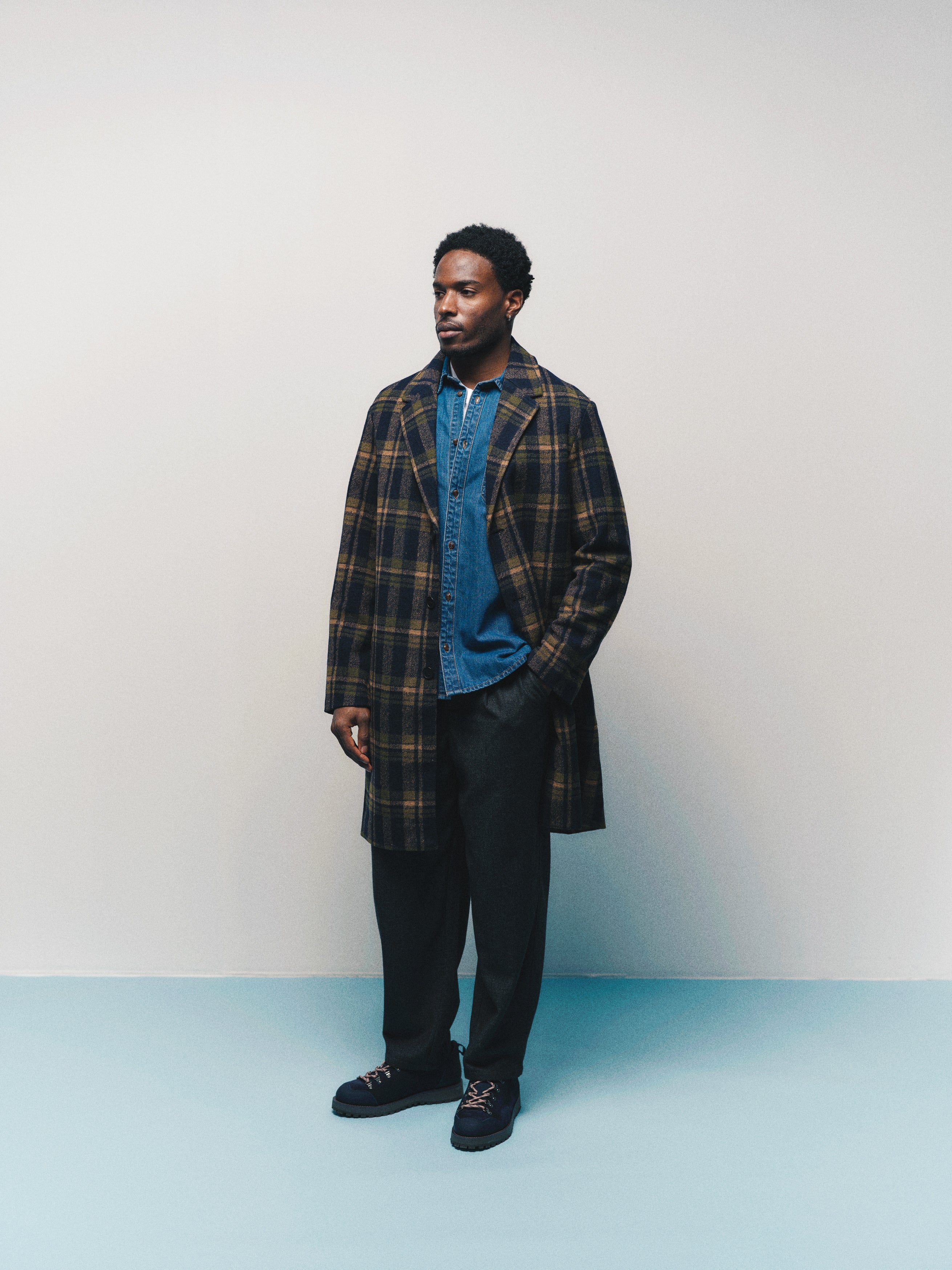 A man stands against a plain background wearing a blue denim shirt, a wool-blend twill dark plaid overcoat, the Gabba GABdavid Twilight Pant in Dark Grey with a loose tapered fit, and black shoes. He looks slightly to the side with a neutral expression.