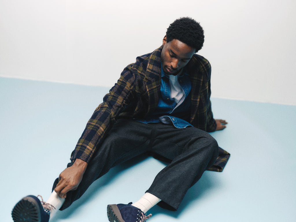 A man in layered clothing and the Gabba GABleeds Chezy Coat in Forest Night sits on a light blue floor, one leg extended and the other bent, looking downward as he adjusts his pant leg near his shoe. This timeless outerwear from Gabba adds sophistication to his tailored silhouette.