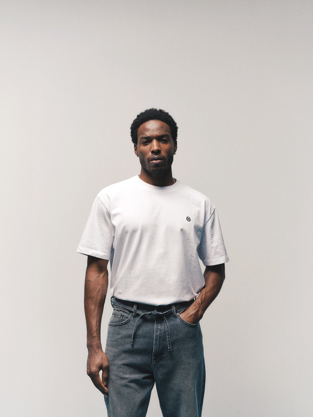 A man stands against a plain, light background wearing the Gabba GABdune Element GOTS SS T-Shirt in white—a classic fit tee made from organic cotton with a small logo—paired with belted gray jeans, looking directly at the camera with a neutral expression.