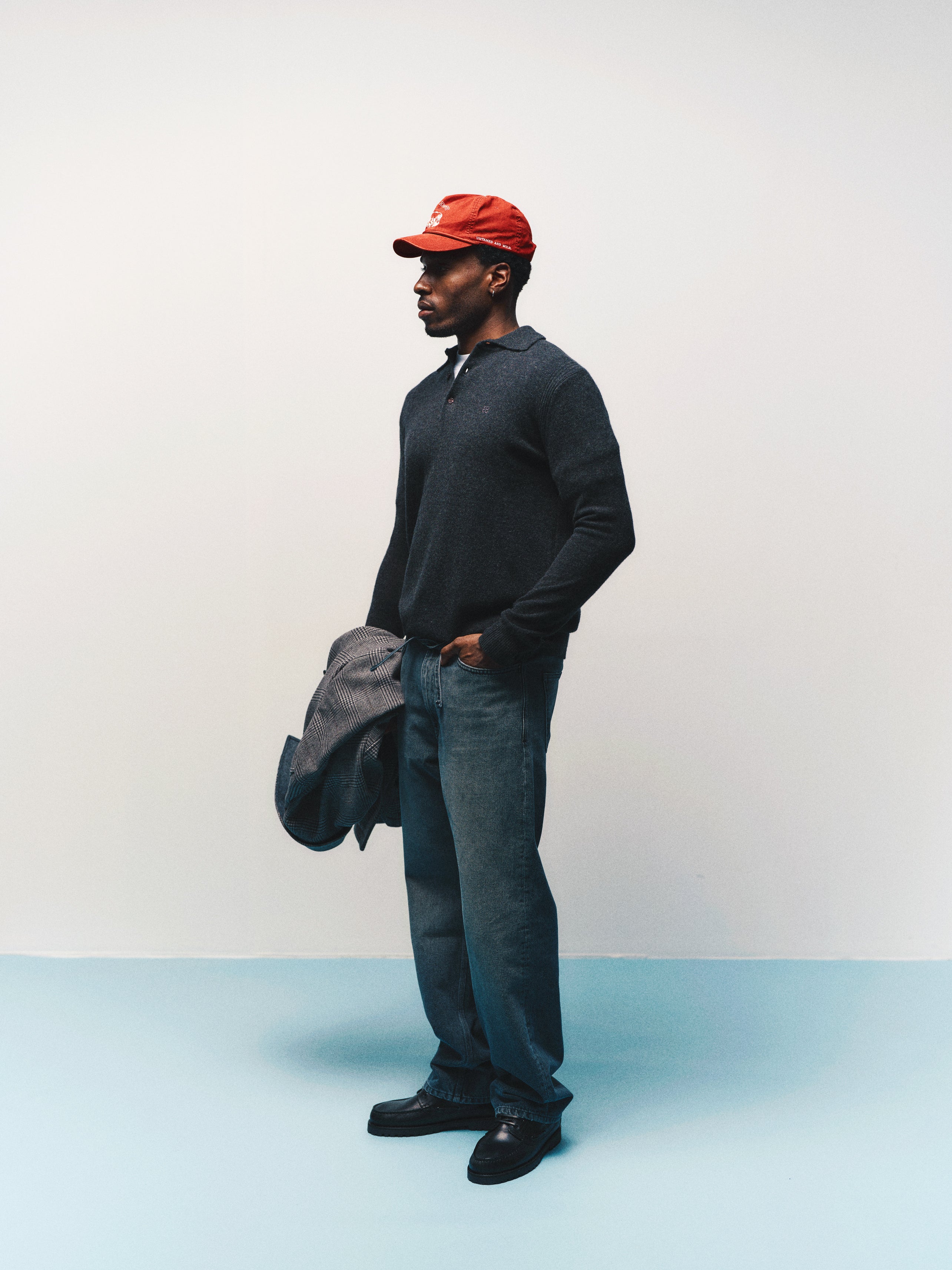 A man in a red cap, jeans, and the Gabba GABworren Polo Aico LS Knit in Dark Grey Mel. stands holding a gray jacket against a plain white and blue background.