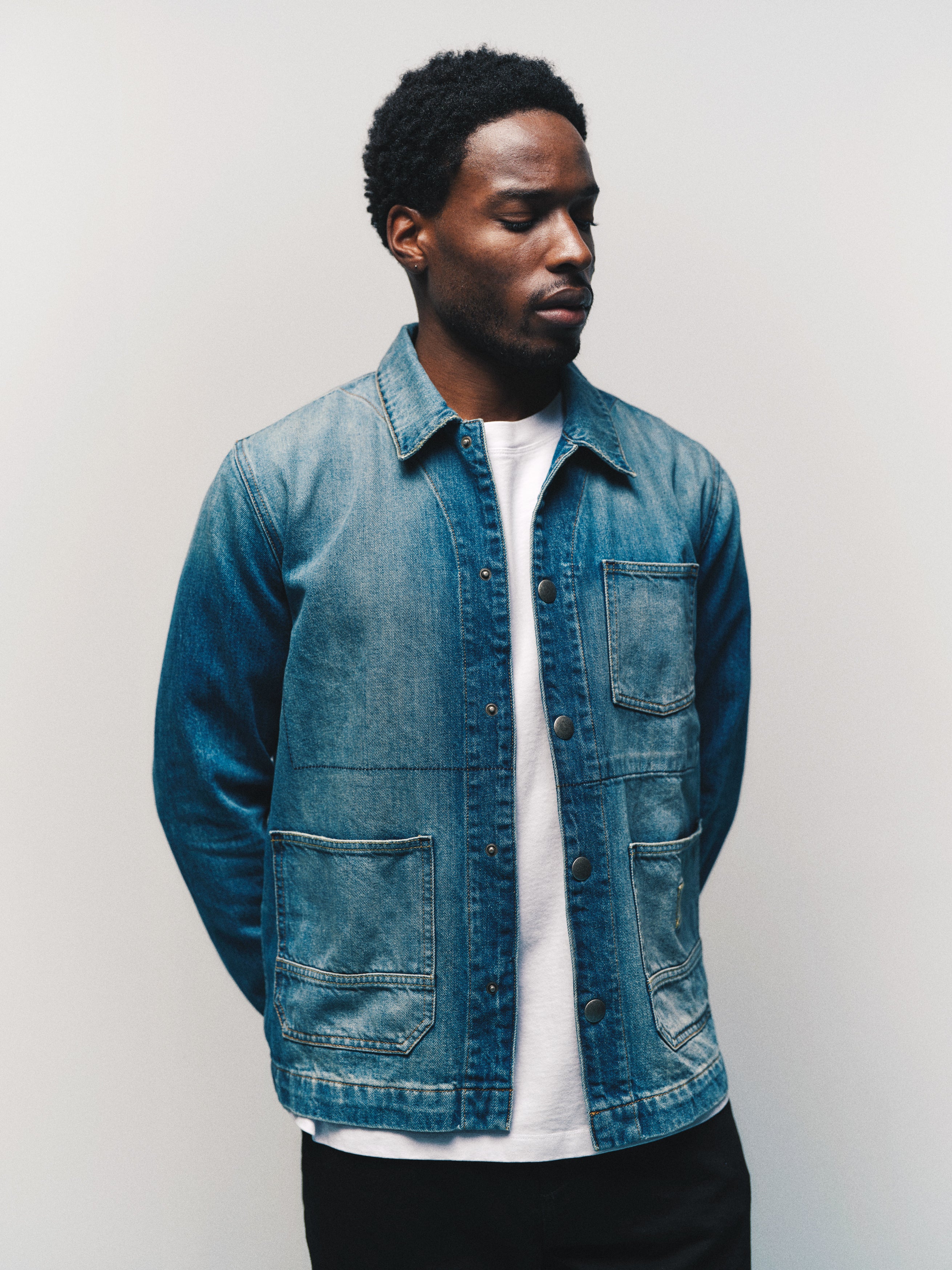 A man stands against a plain light-colored background with his hands behind his back, looking slightly down and to the side, wearing the Gabba GABbeta Wolv DS0004 LS Overshirt in dark blue denim over a white t-shirt.