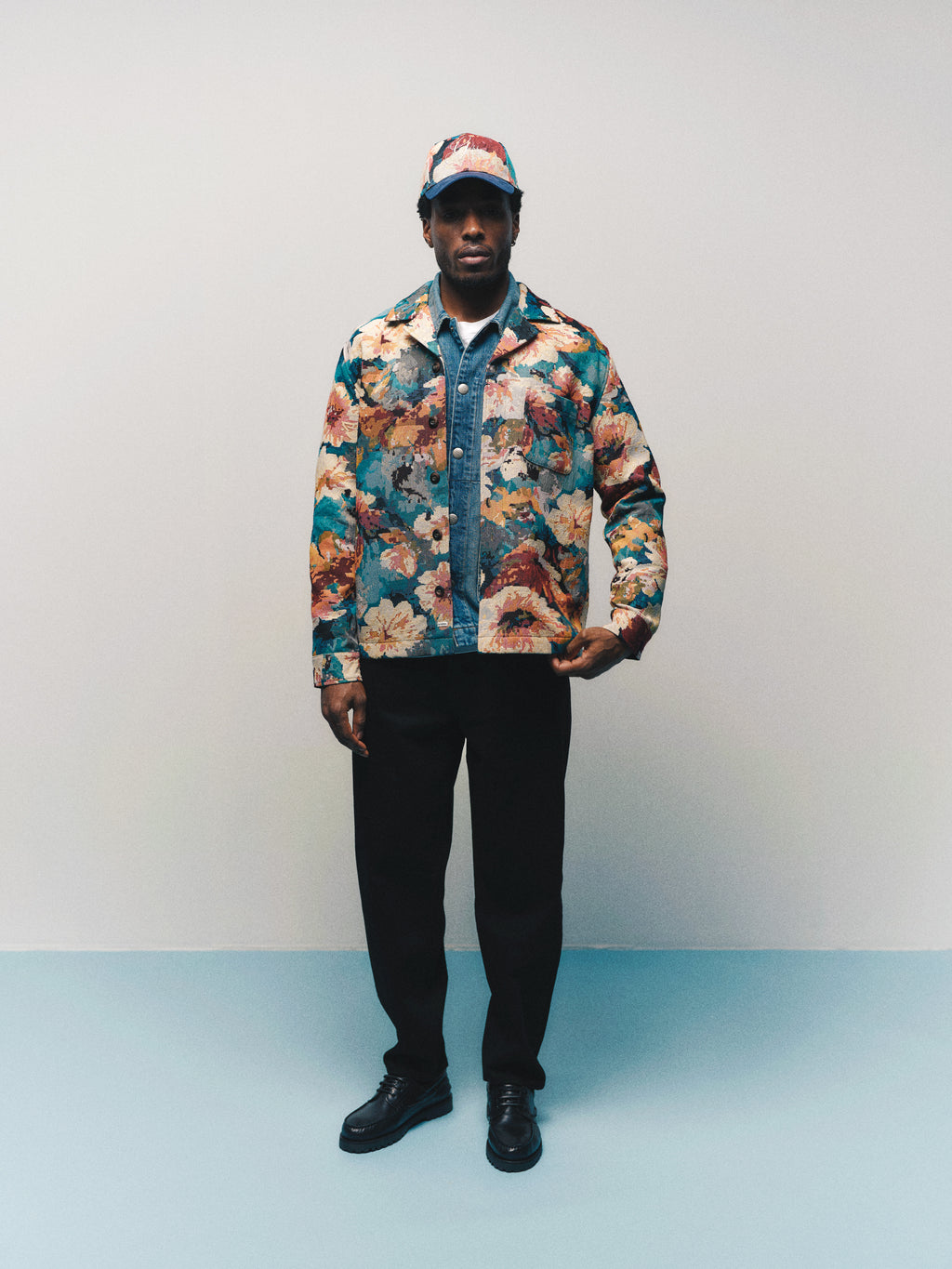 A man stands against a plain background wearing the Gabba GABsimone Reso Fiore LS Overshirt in a multi pattern, paired with a matching cap, a blue floral jacquard relaxed-fit resort collar shirt, black pants, and black shoes, facing the camera with a neutral expression.