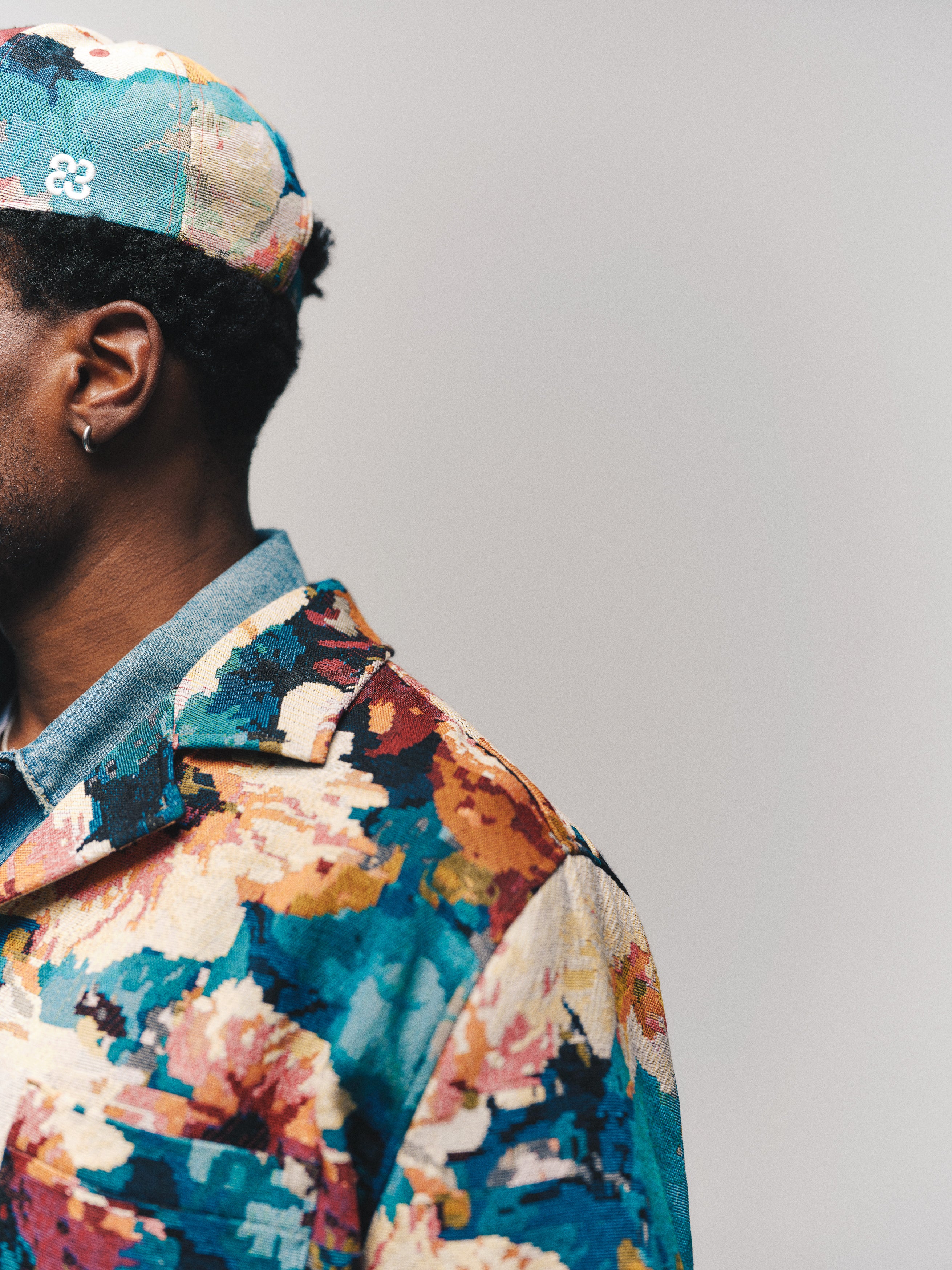 A person wearing the Gabba GABsimone Reso Fiore LS Overshirt in a multi-patterned design and a matching cap, shown from the shoulders up, facing left against a plain light background.