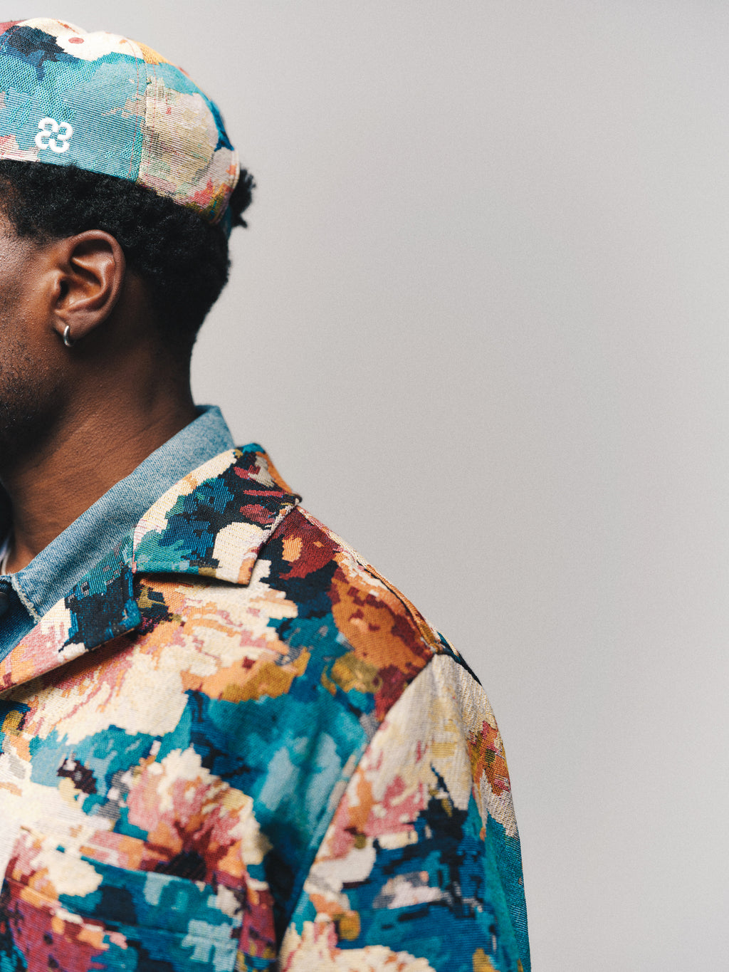 A person wearing the Gabba GABsimone Reso Fiore LS Overshirt in a multi-patterned design and a matching cap, shown from the shoulders up, facing left against a plain light background.