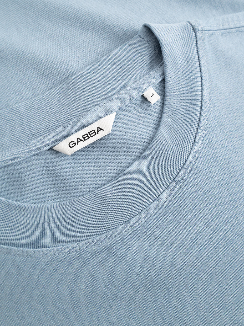 Close-up of the GABlegin Wix Boxy Hour SS T-shirt in Dusty Blue by Gabba, crafted from garment-washed cotton. The relaxed, boxy fit is highlighted as it’s laid flat on a matching light blue background, featuring a white Gabba label and an L size tag. A subtle embroidered Gabba logo adds a refined touch to this effortlessly modern piece.