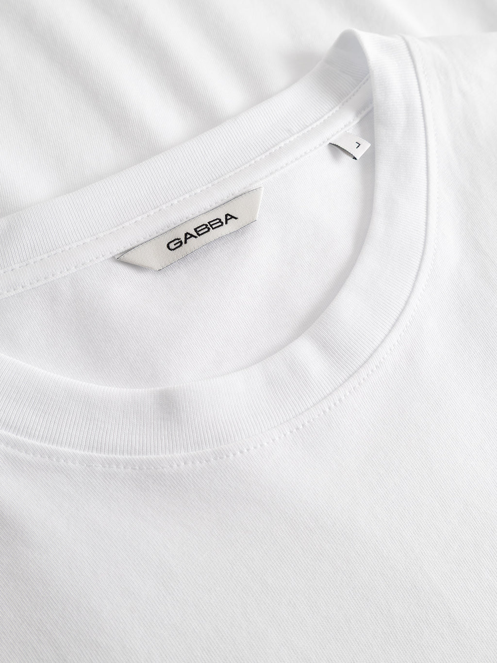 Close-up of two stacked white Gabba GABdune Element GOTS SS T-Shirts in classic fit, with the top shirt’s tag showing the Gabba brand and a size 1 label, neatly arranged on a flat surface.