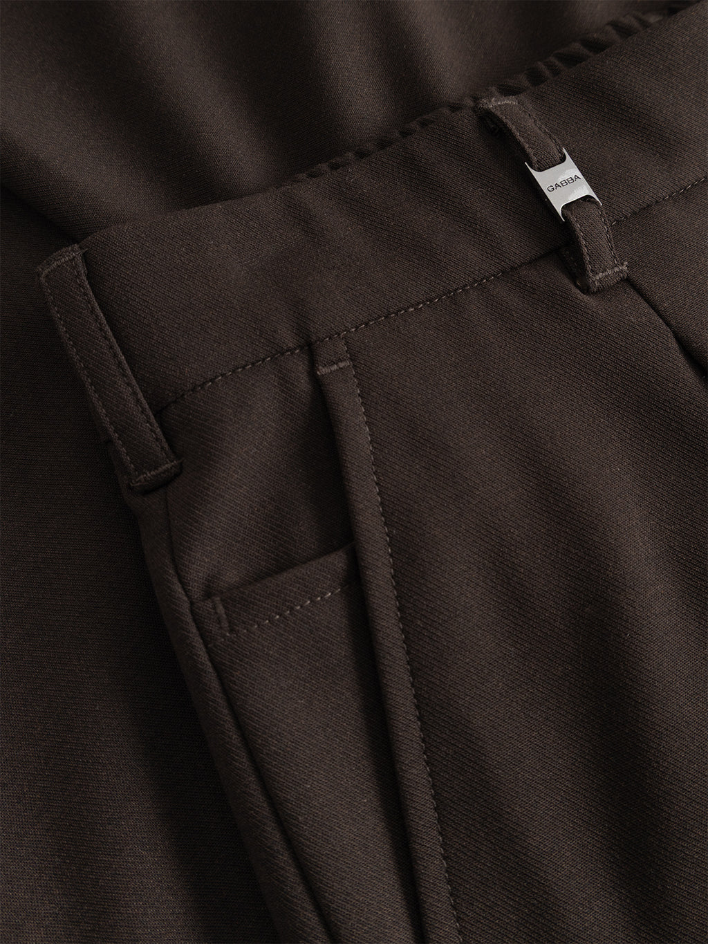 Close-up of Gabba’s GABnapoli 19 Phin GRS - Brown Slate tailored dress pants, crafted from a smooth wool blend chinos fabric with recycled polyester. These dark brown slate trousers feature a regular straight fit, belt loops, pleats, and a waistband tab adorned with a metal brand tag. Part of the matching jacket is visible in the background.