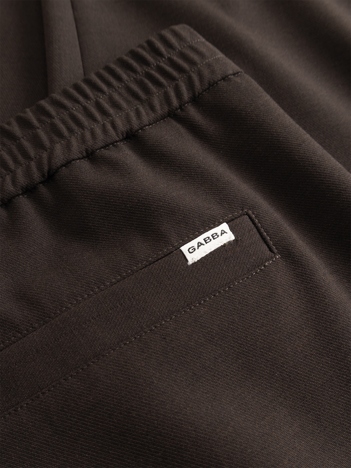 Close-up of the GABnapoli 19 Phin GRS - Brown Slate chinos by Gabba, showcasing a dark brown wool blend with an elastic waistband, back pocket, and a small white tag bearing the Gabba logo sewn beside the pocket seam. Designed in a regular straight fit and crafted with recycled polyester for enhanced comfort and sustainability.