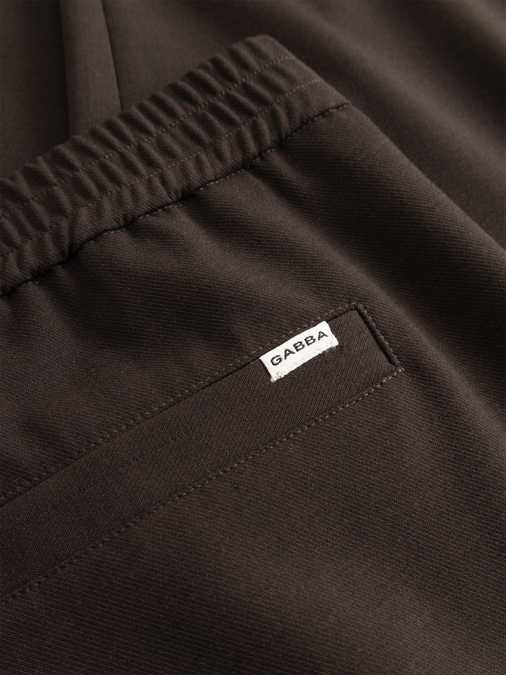 Close-up of the GABnapoli 19 Phin GRS - Brown Slate chinos by Gabba, showcasing a dark brown wool blend with an elastic waistband, back pocket, and a small white tag bearing the Gabba logo sewn beside the pocket seam. Designed in a regular straight fit and crafted with recycled polyester for enhanced comfort and sustainability.